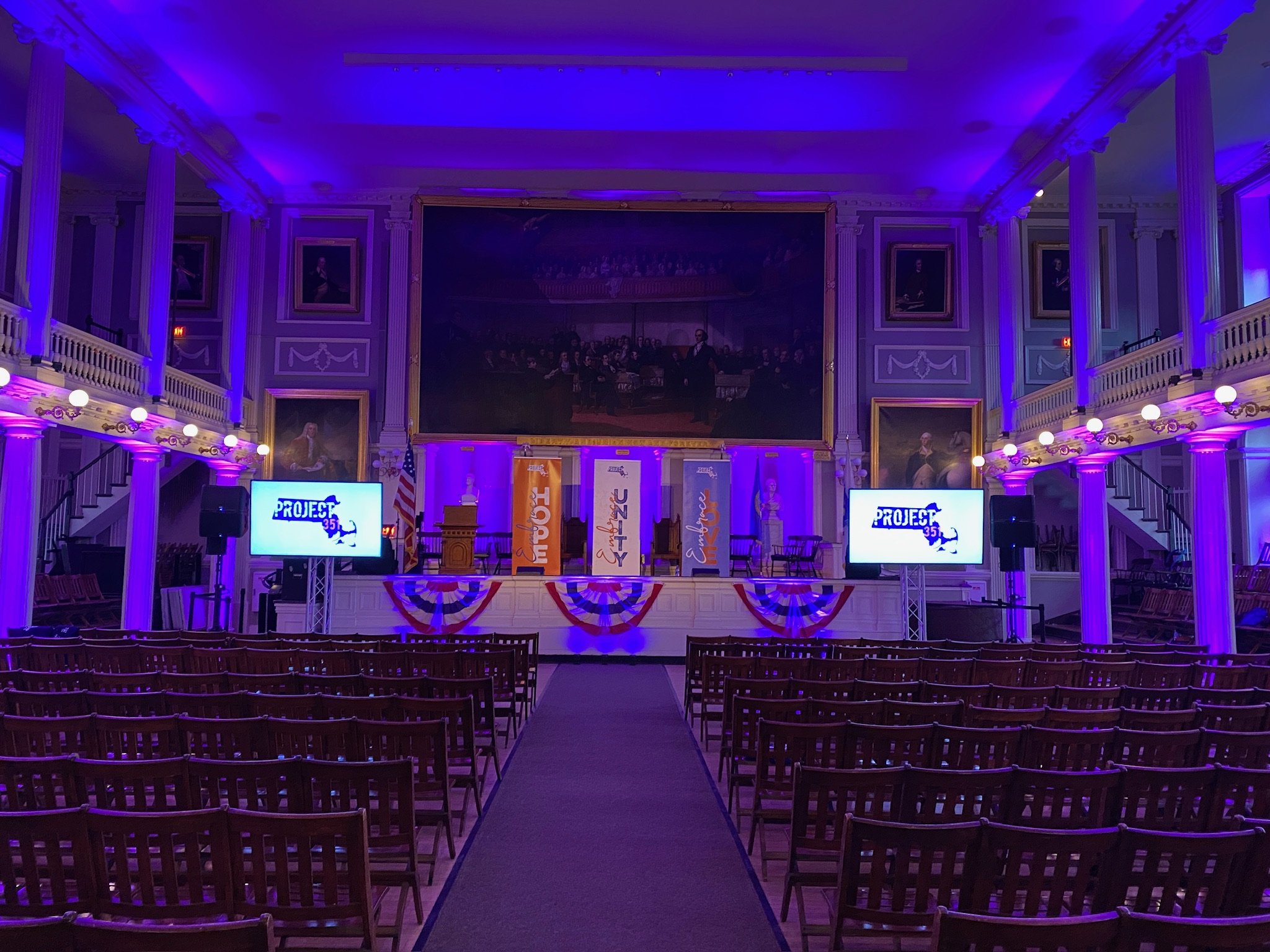A decorated event hall with purple lighting, banners, and chairs arranged facing a stage with two screens displaying the text 'PROJECT' and a graphic of the United States map, set for a formal presentation or celebration.