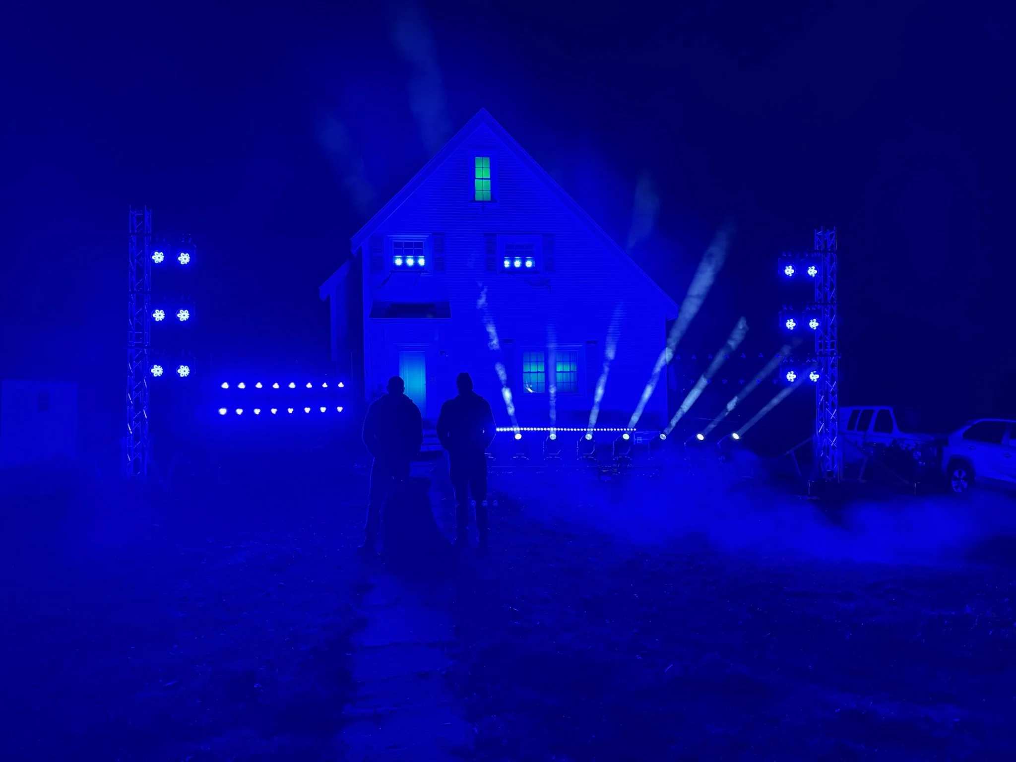 Nighttime outdoor scene with a house illuminated by blue lights, and two people standing in front of it. There is stage lighting equipment on both sides, and some parked vehicles are visible in the background.