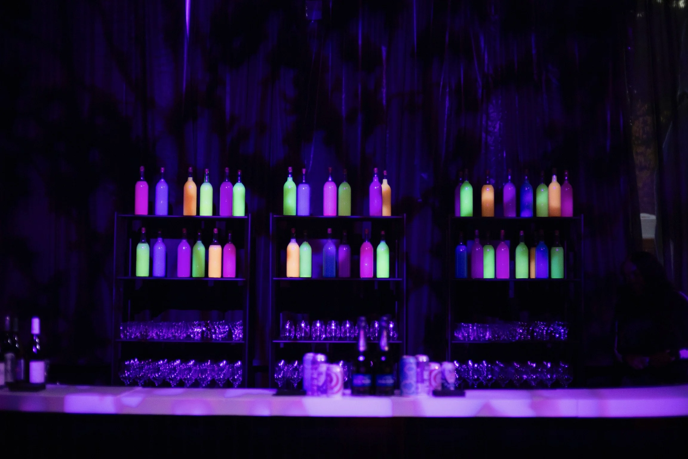 Display of colorful bottles and glasses illuminated under purple lighting at a bar.