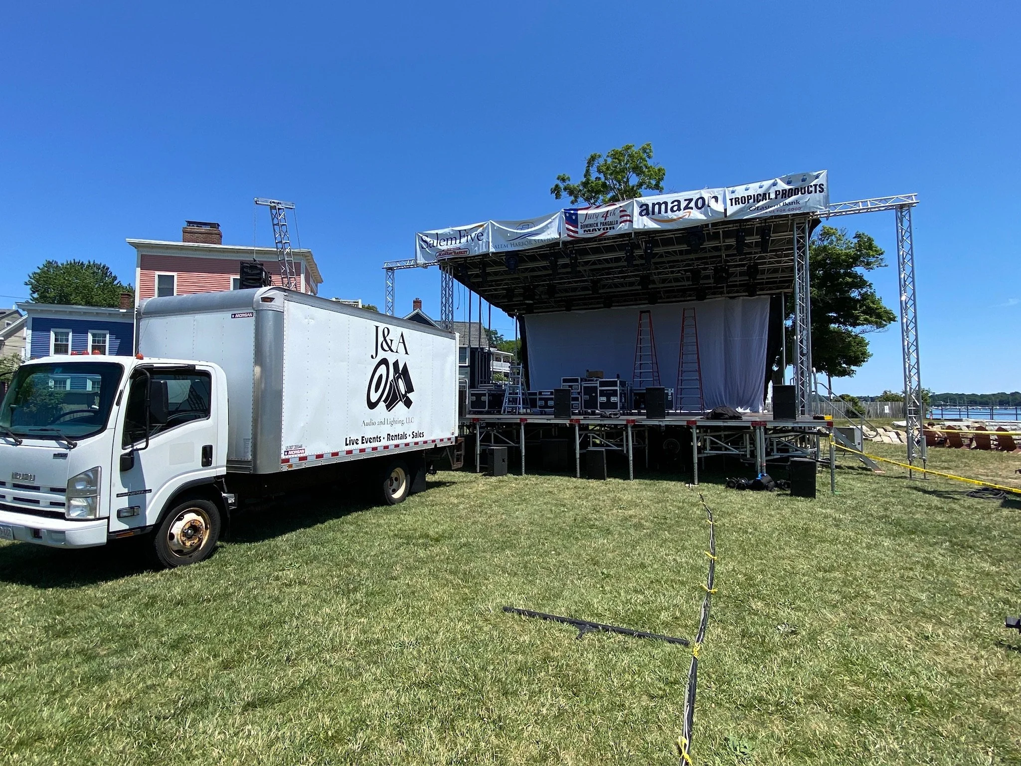 An outdoor event setup with a stage; a truck labeled J&A Audio and Lighting LLC parked next to it; banners overhead on the stage display Amazon, Tropical Products, and Salem Five; the sky is clear and blue, and there are houses and trees in the backg