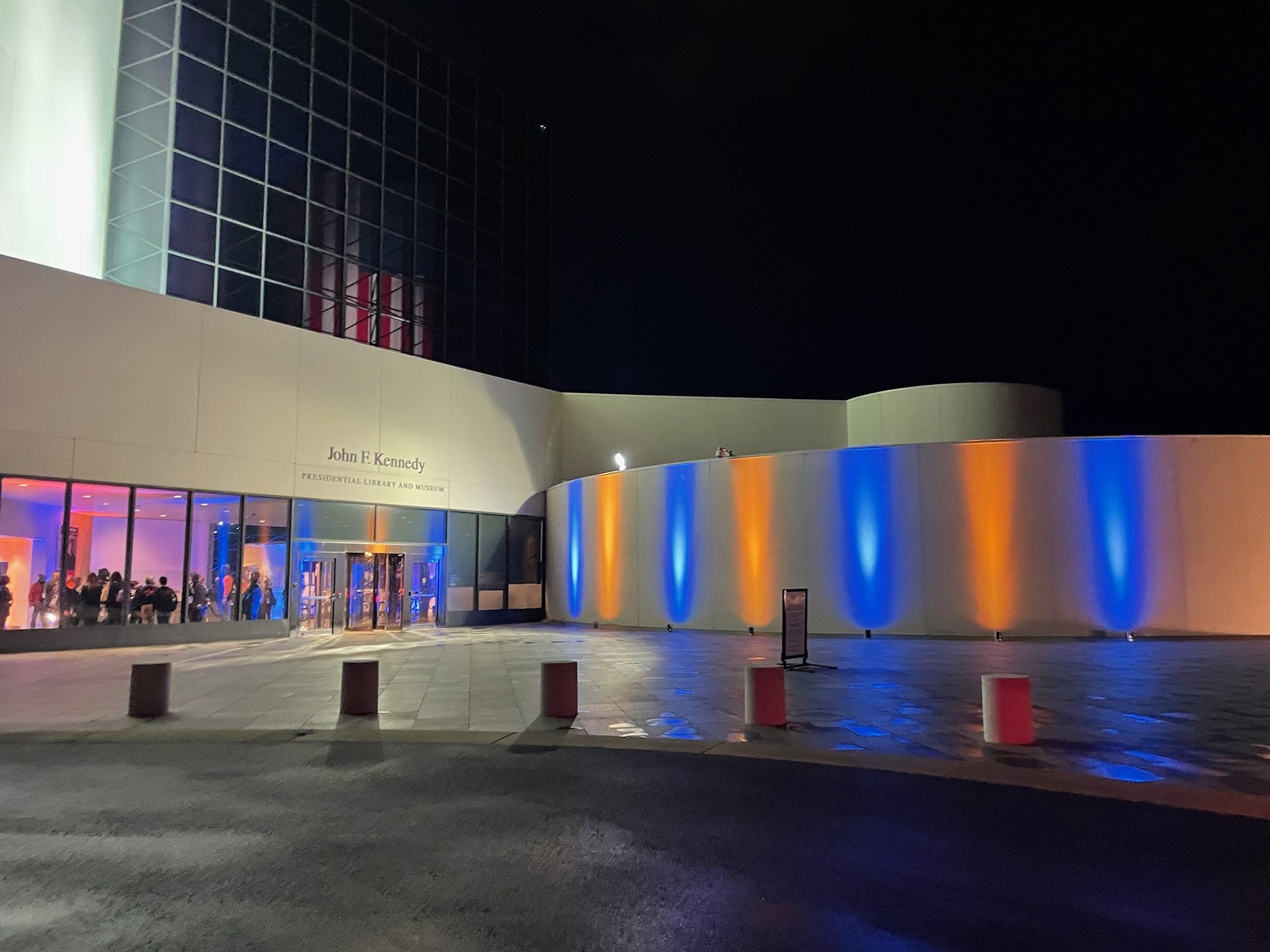 Night view of the John F. Kennedy Presidential Library and Museum, with colorful blue and orange lighting illuminating the building's exterior, and people visible inside through glass windows.