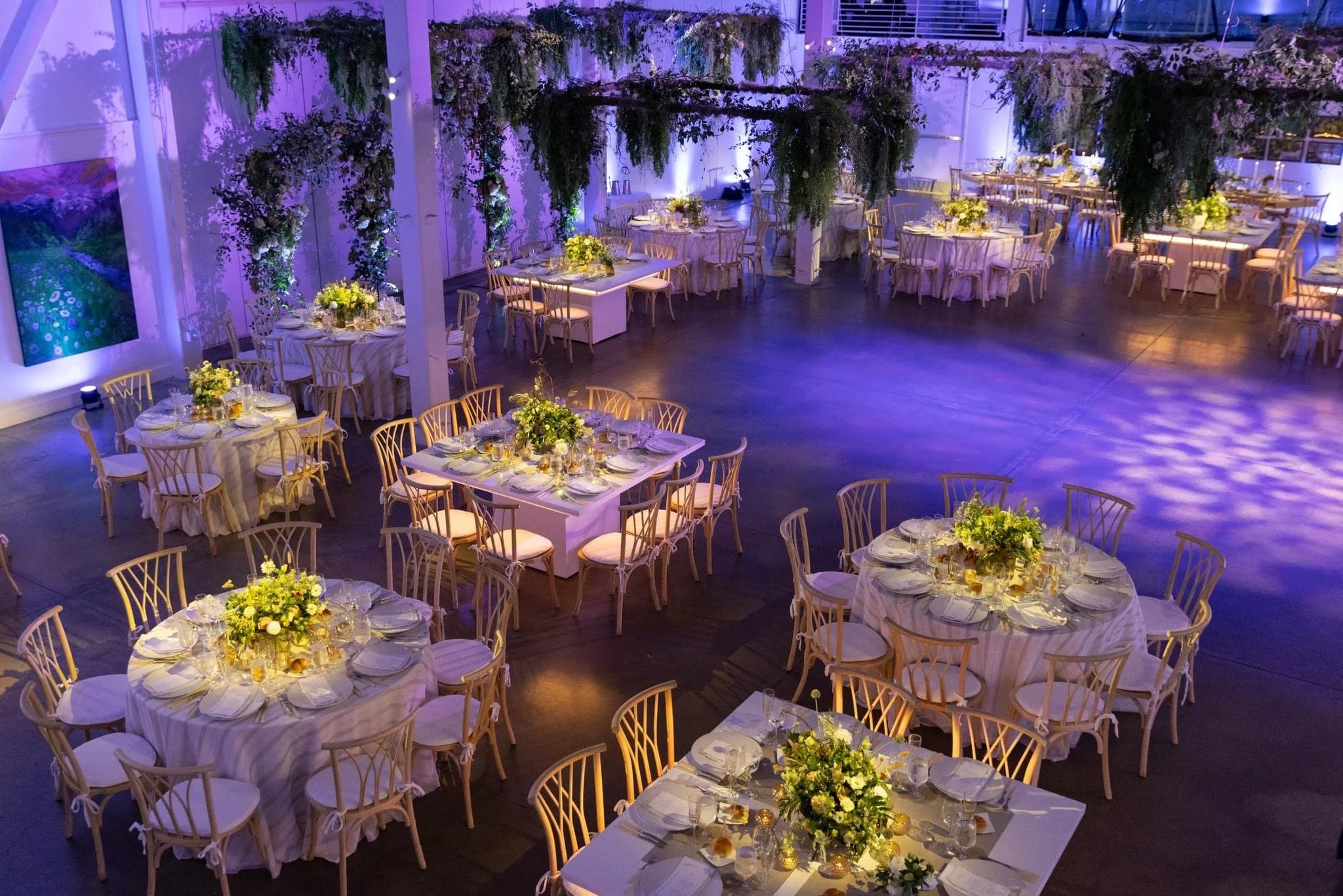 Elegant event space decorated with round and rectangular tables with white tablecloths, topped with floral centerpieces, set for a formal dinner. Purple and blue lighting highlights hanging greenery and floral arrangements.
