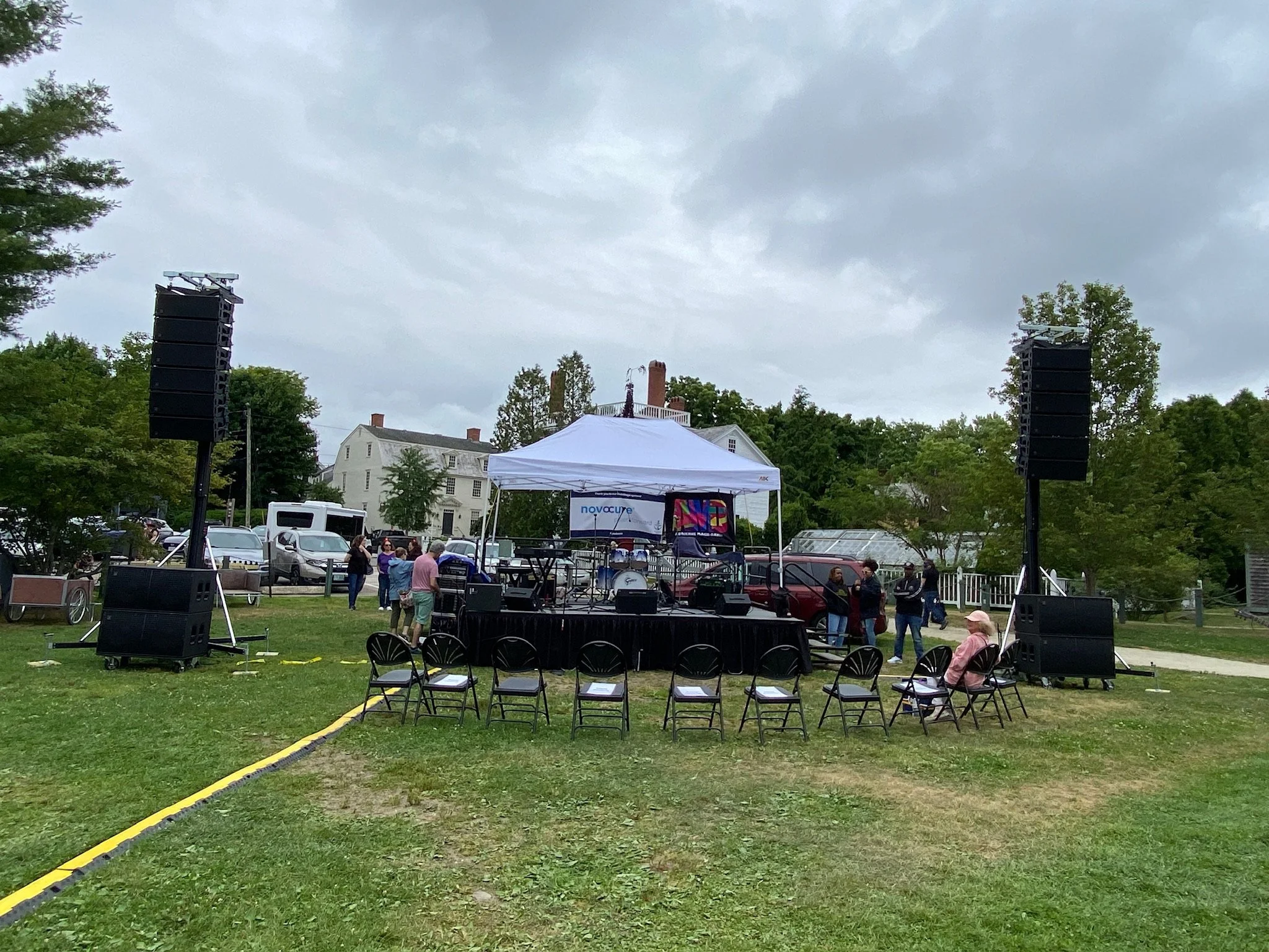 Outdoor stage setup with speakers, chairs, and a canopy tent, surrounded by trees and parked cars, under cloudy sky.
