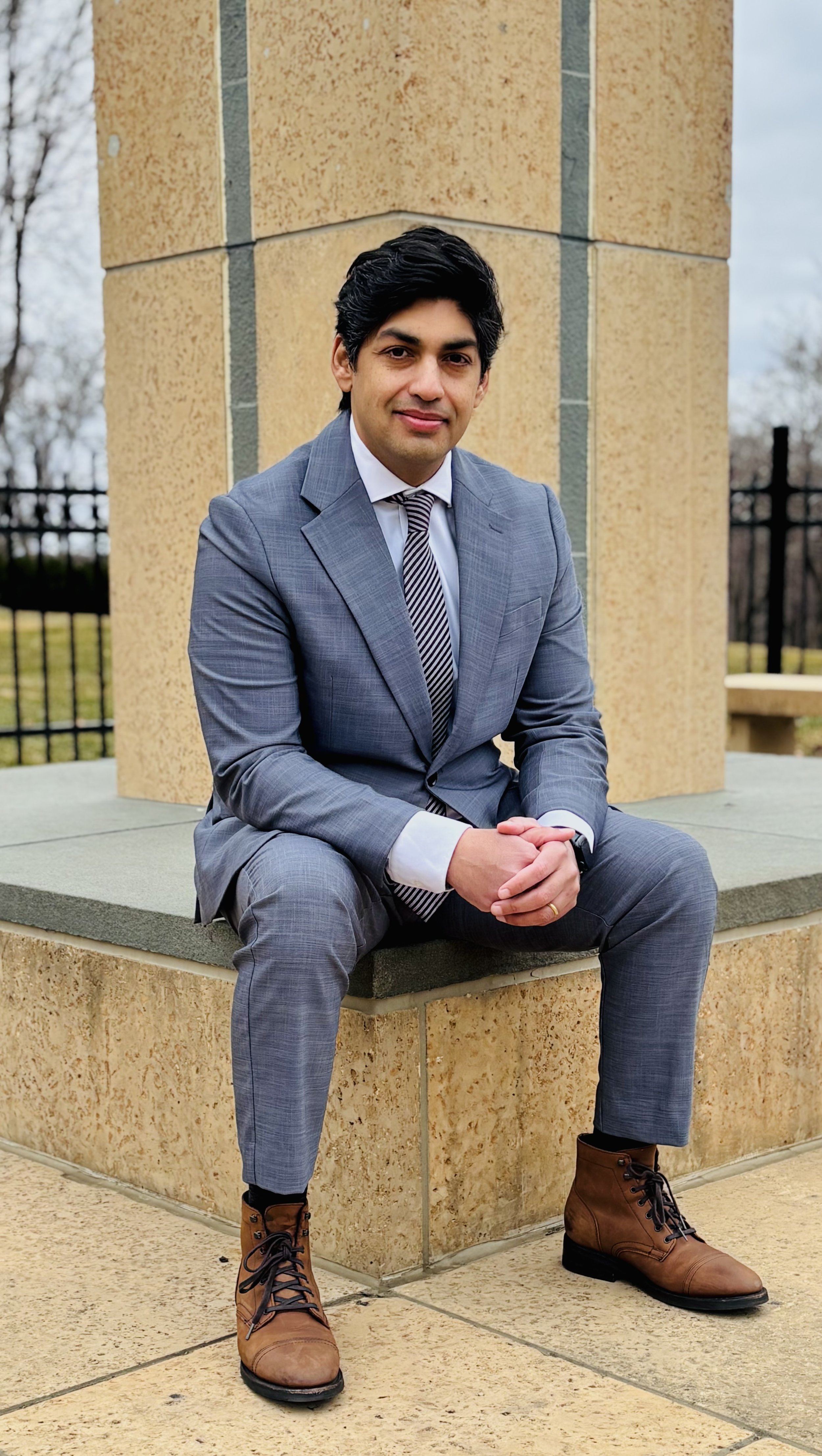 Man in a gray suit seated in front of a pillar. His hands are clasped, he leans forward, and he is smiling.