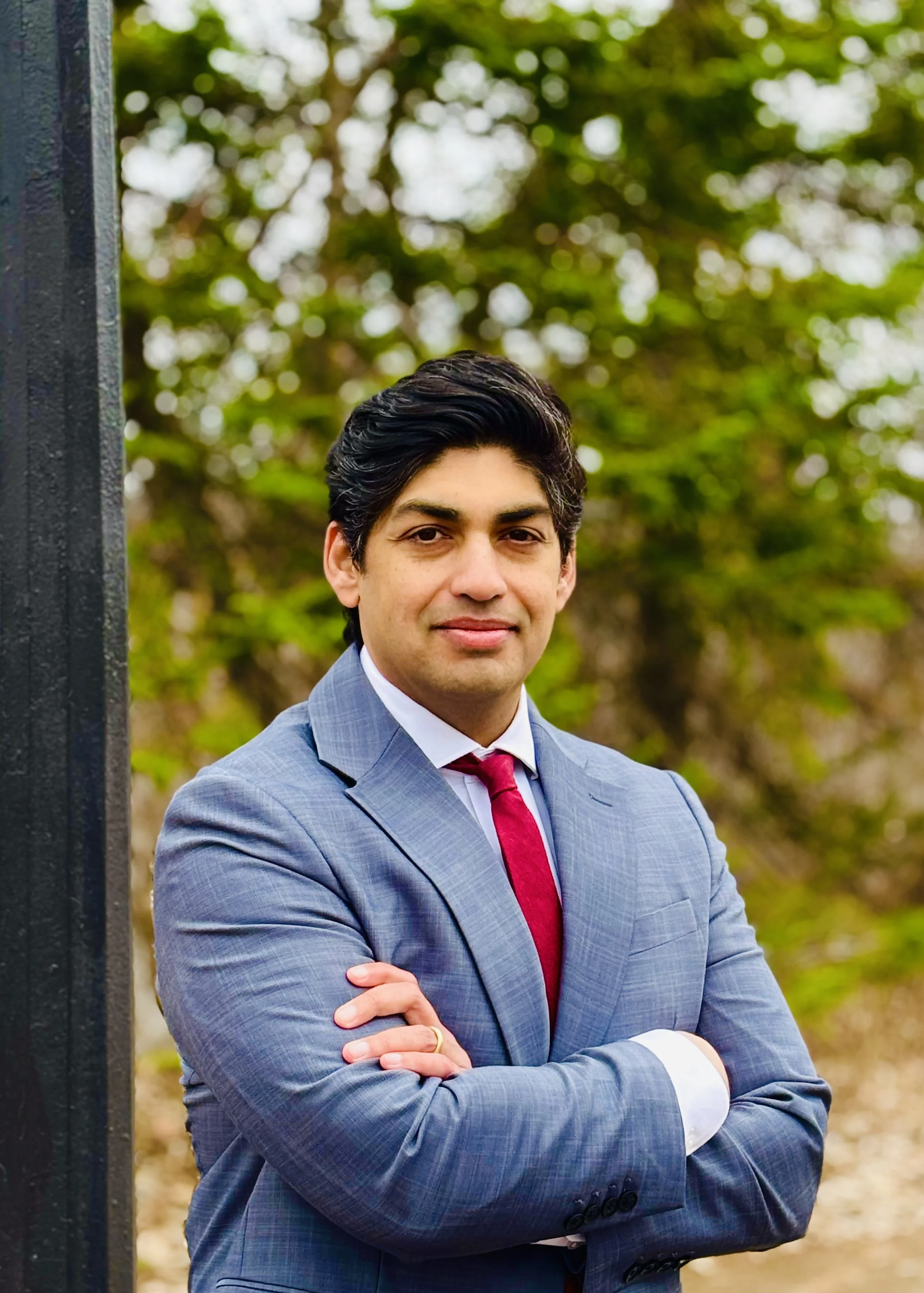Man in a gray suit standing next to a pole with his arms crossed. He looks at the camera confidently.