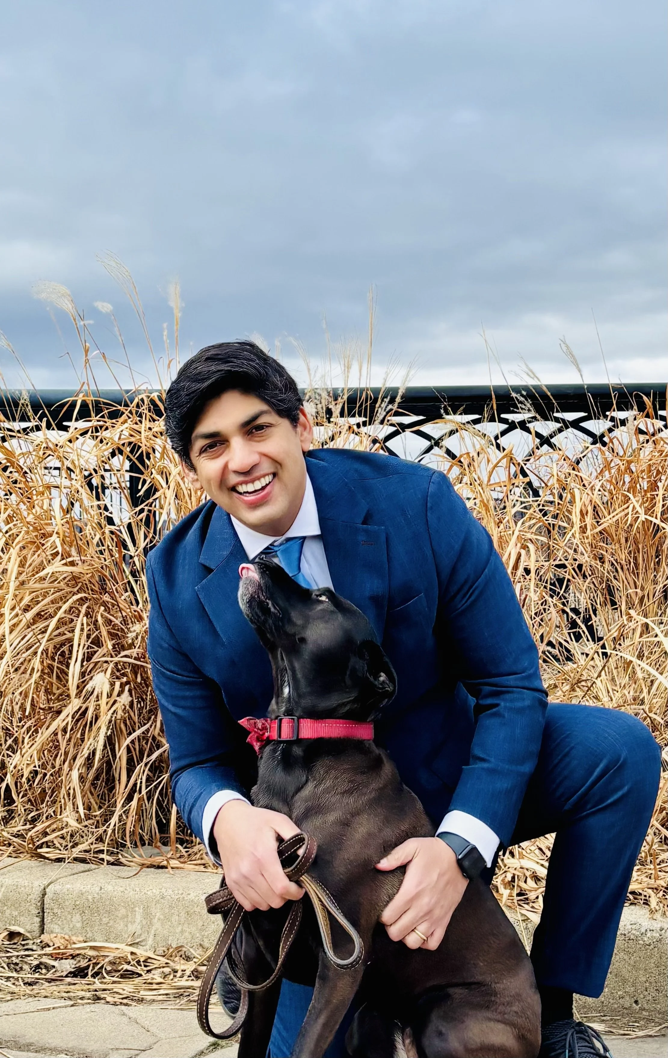 A man in a blue suit kneeling down behind a black dog. The dog is looking up the man, about to lick him. The man is scratching the dog's fur and looking at the camera, laughing.
