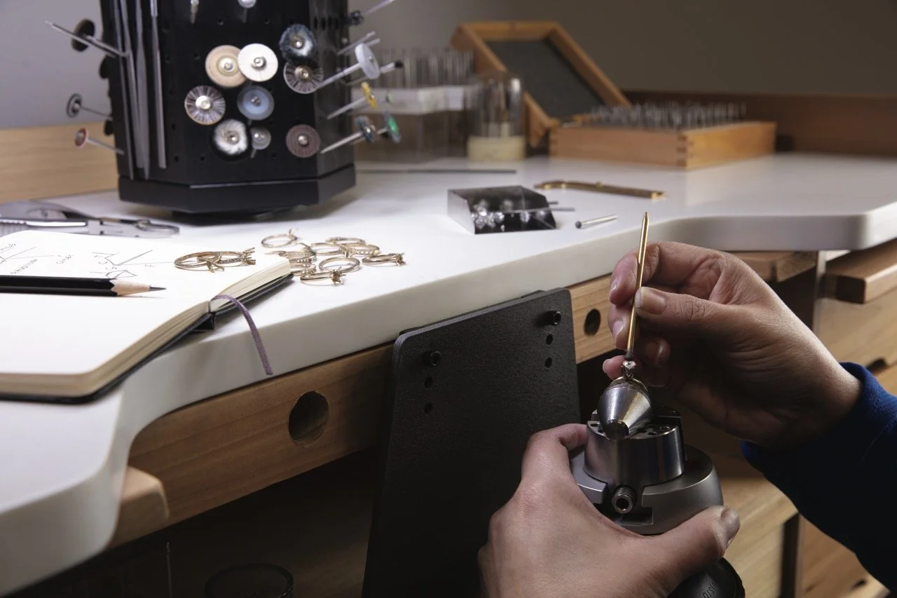 A jeweler working at a workbench, using a tool to engrave or polish a piece of jewelry.