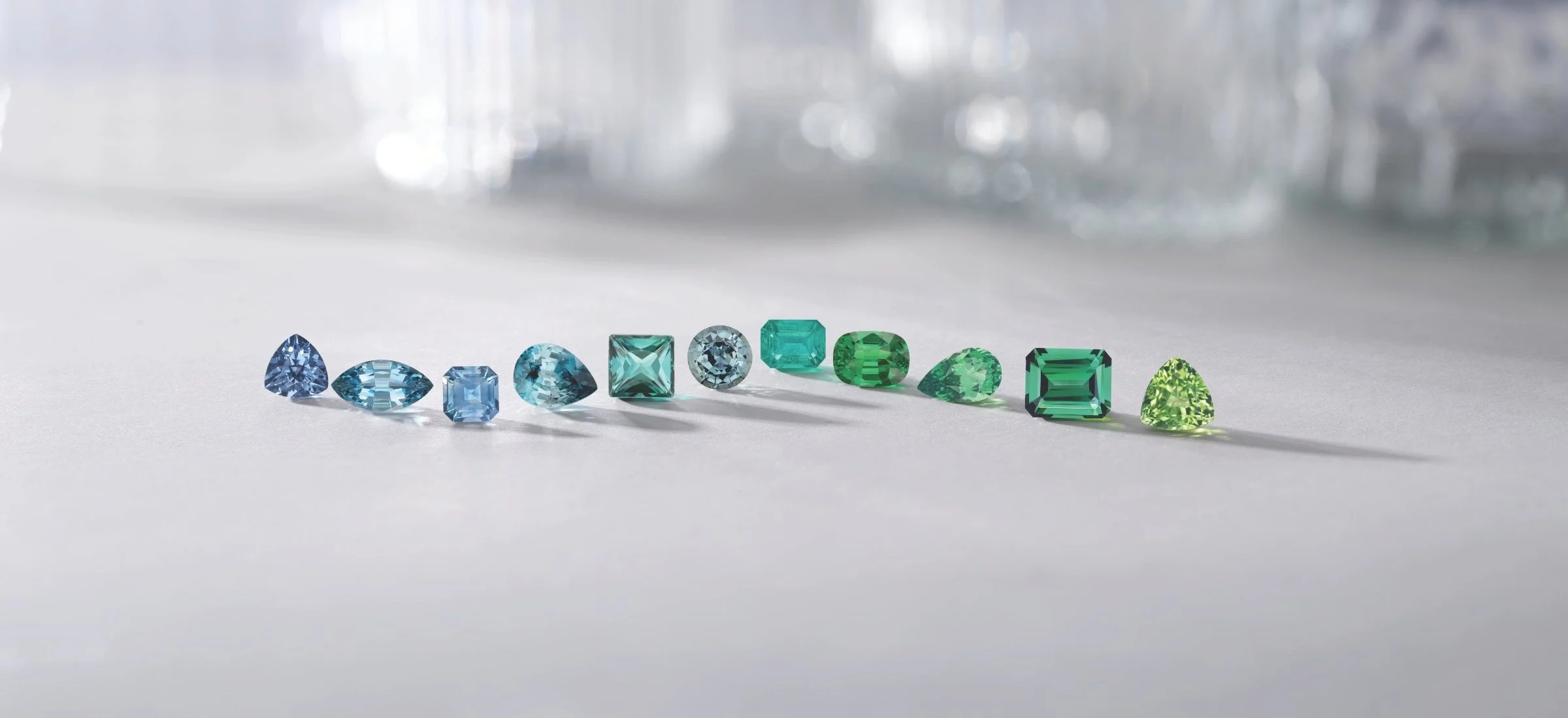 A row of blue and green loose gemstones in varied cuts arranged on a soft gray surface.