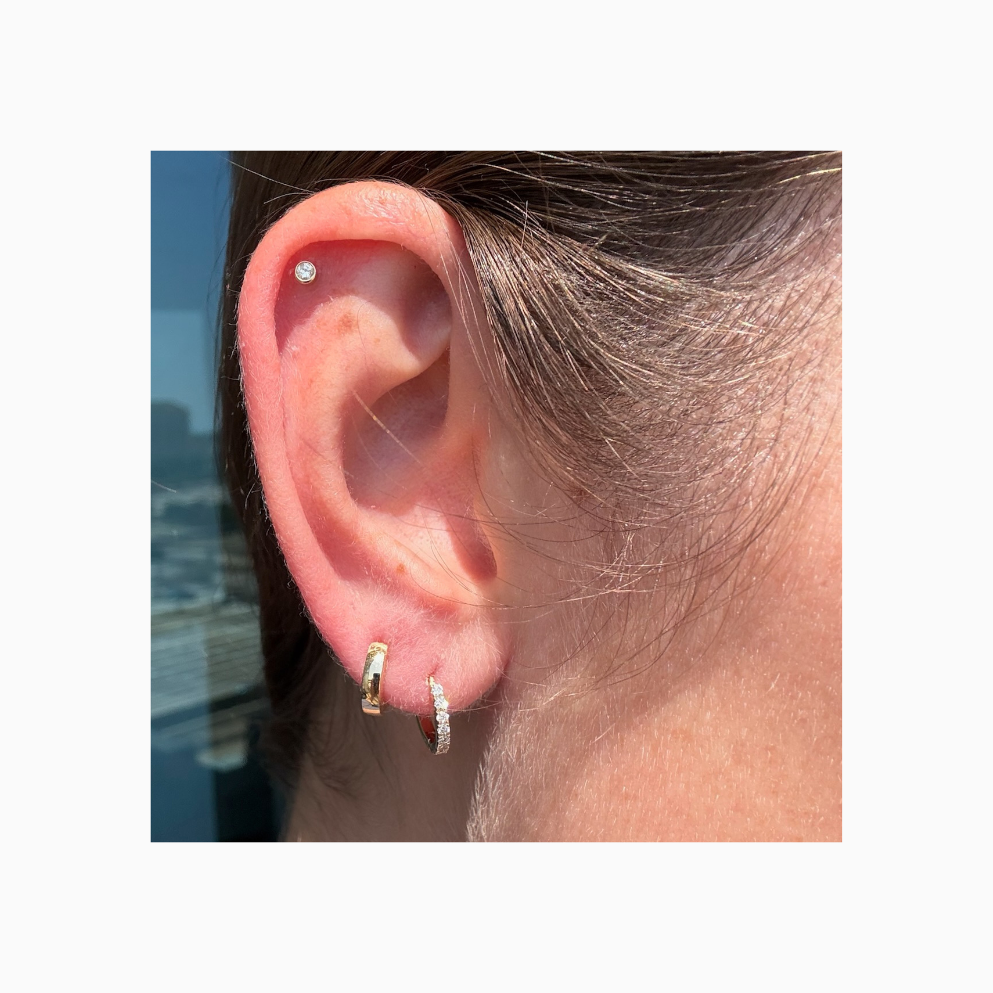 14K yellow gold huggie hoop worn on ear showing its petite, close-fitting size. Also pictured diamond yellow gold huggie on ear lobe and diamond stud on ear cartilage