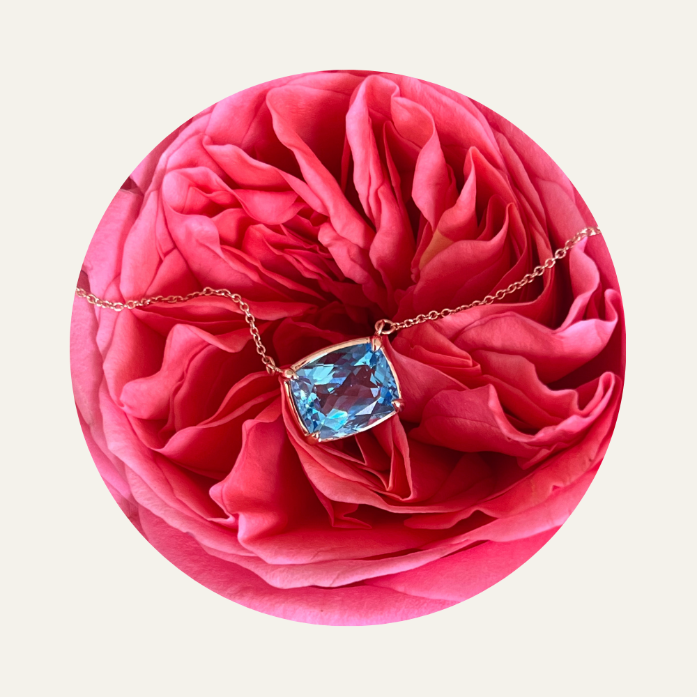 A necklace with a large blue natural topaz in a bezel set pendant resting on a pink rose flower.