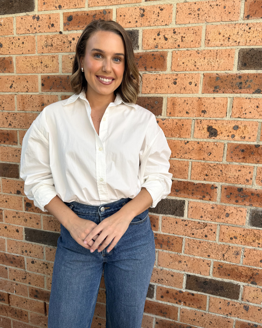A young woman with shoulder-length brown hair, wearing a white button-up shirt and blue jeans, standing against a brick wall, smiling at the camera.