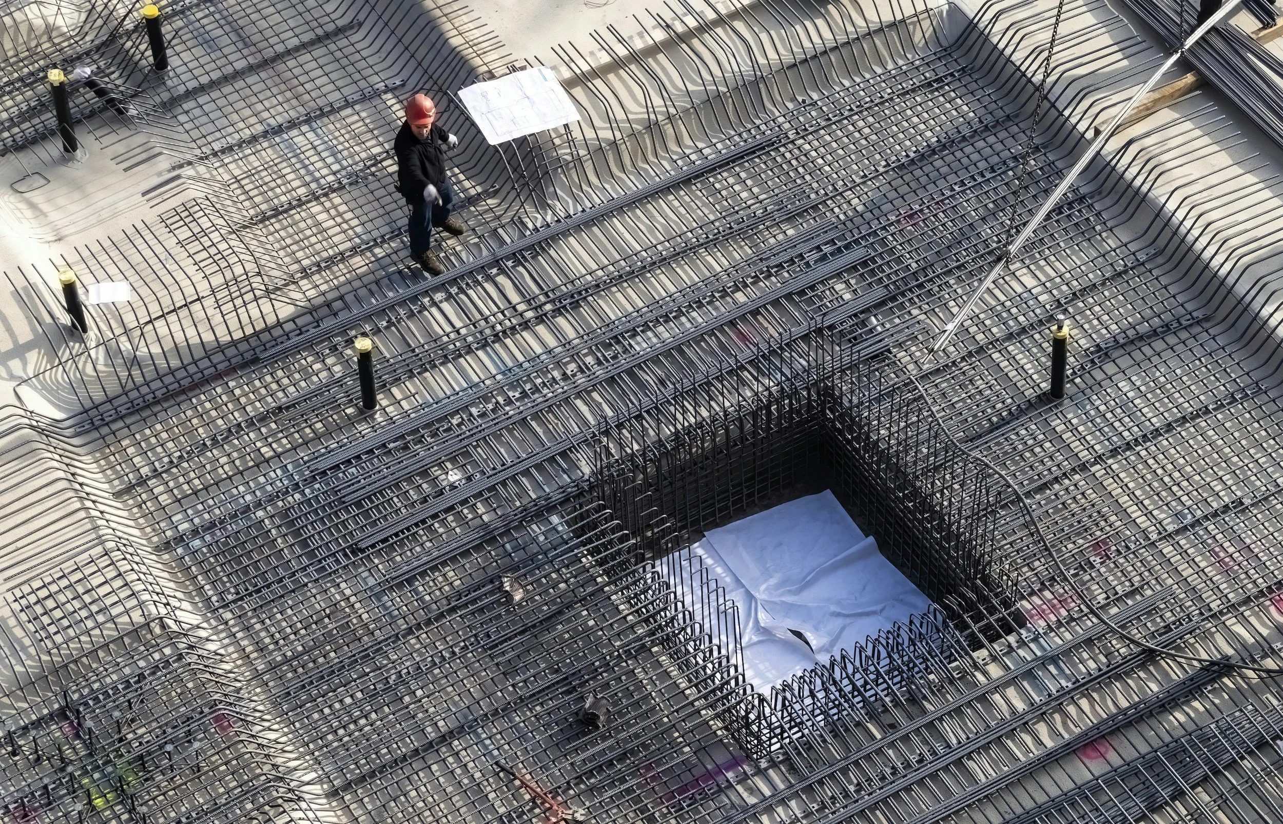 Construction worker standing on steel rebar framework on a building rooftop, wearing a red helmet and dark clothing.