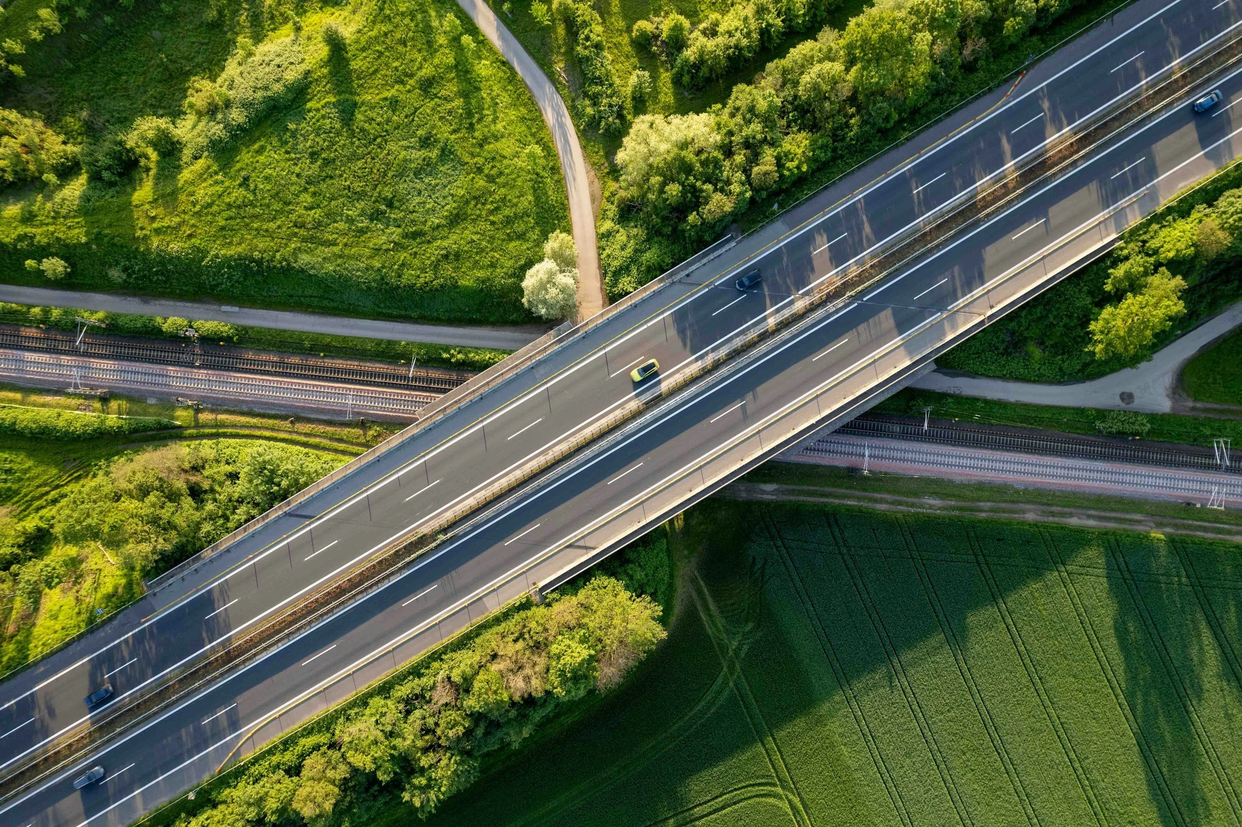 An aerial view of a highway overpass with green fields and trees around it, showing a few cars traveling on the road.