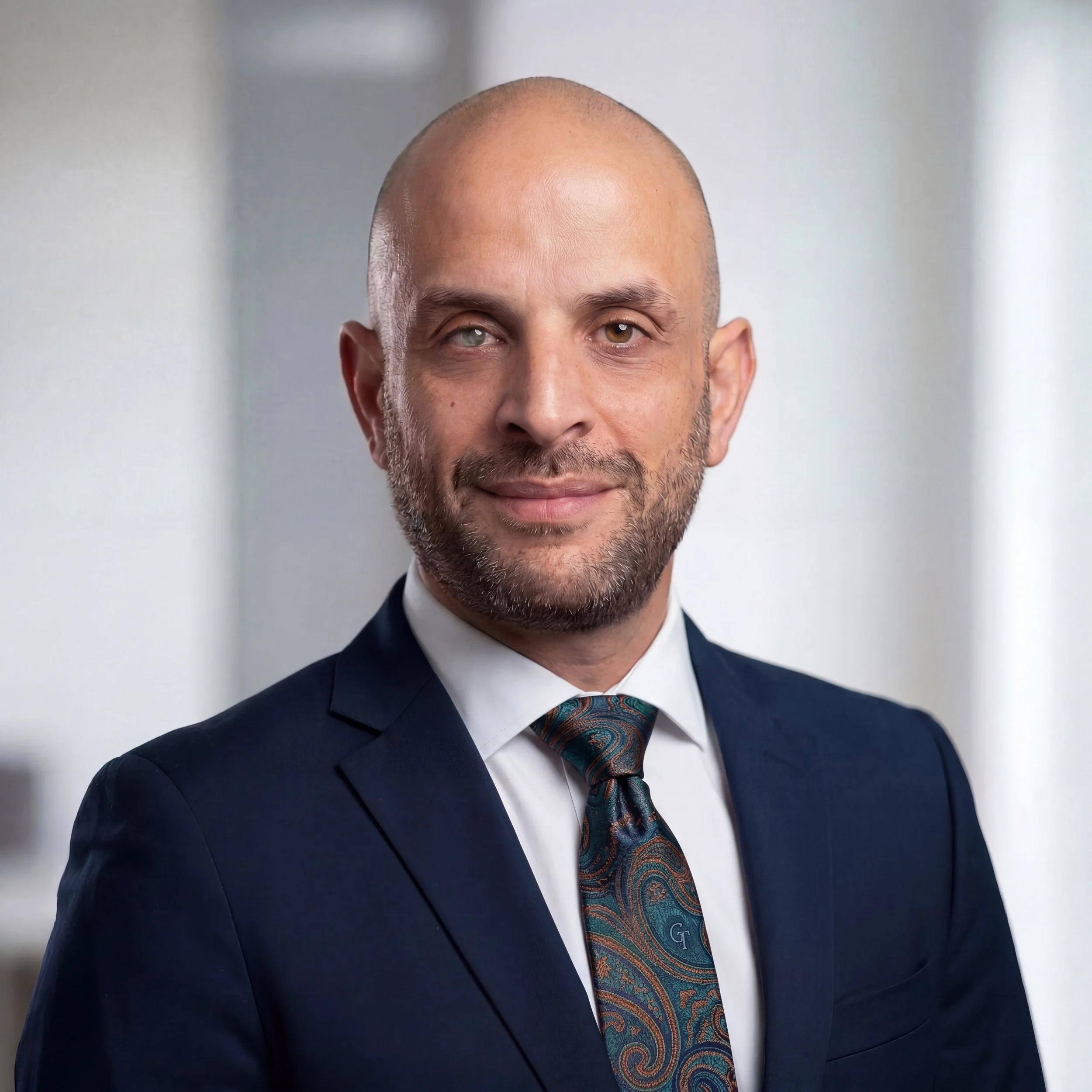A professional man wearing a dark suit, white shirt, and a colorful paisley tie, smiling with a neutral background.