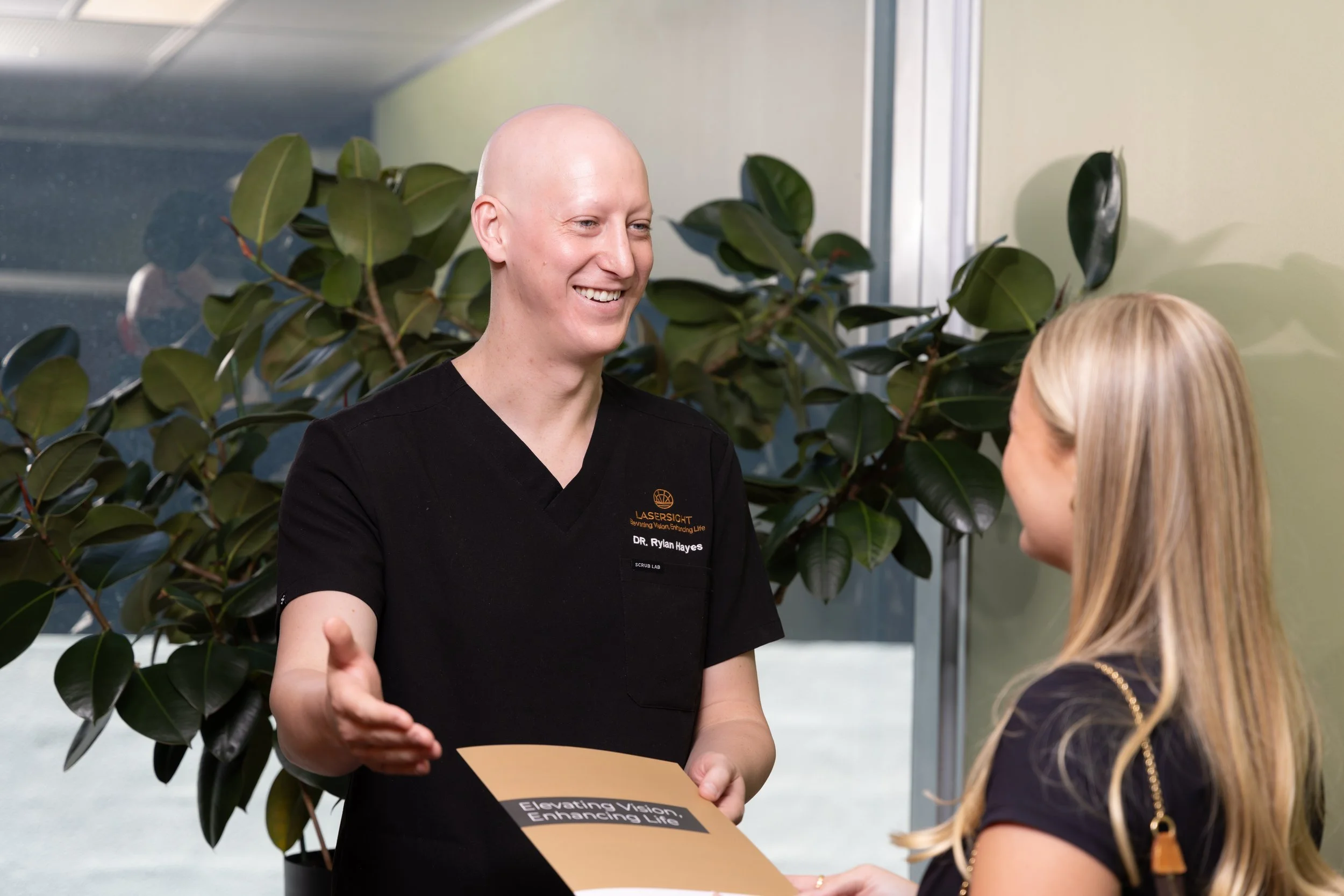 Dr Rylan Hayes in healthcare scrubs discussing pterygium surgery with a young girl in a black shirt in a medical clinic with green plants.