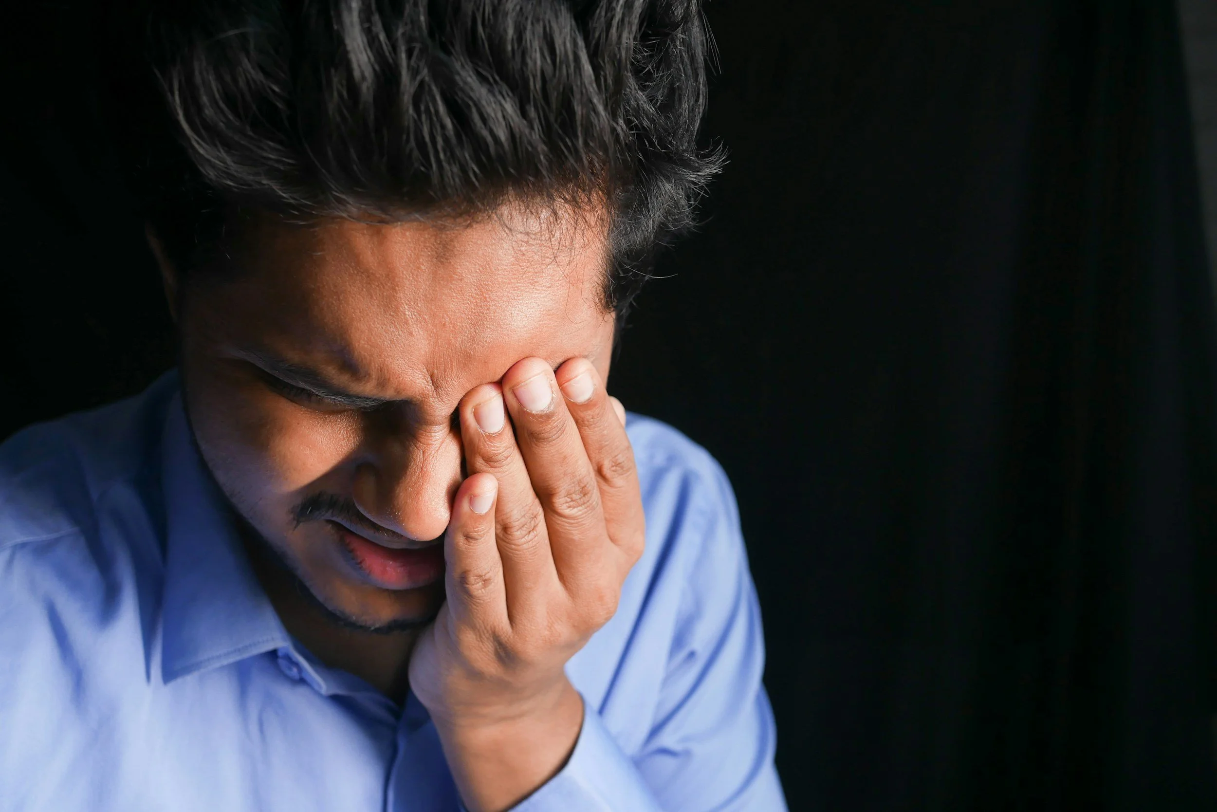 A man with dark hair, wearing a blue shirt, has his face covered with his hands, appearing distressed from the pain of an inflamed pterygium
