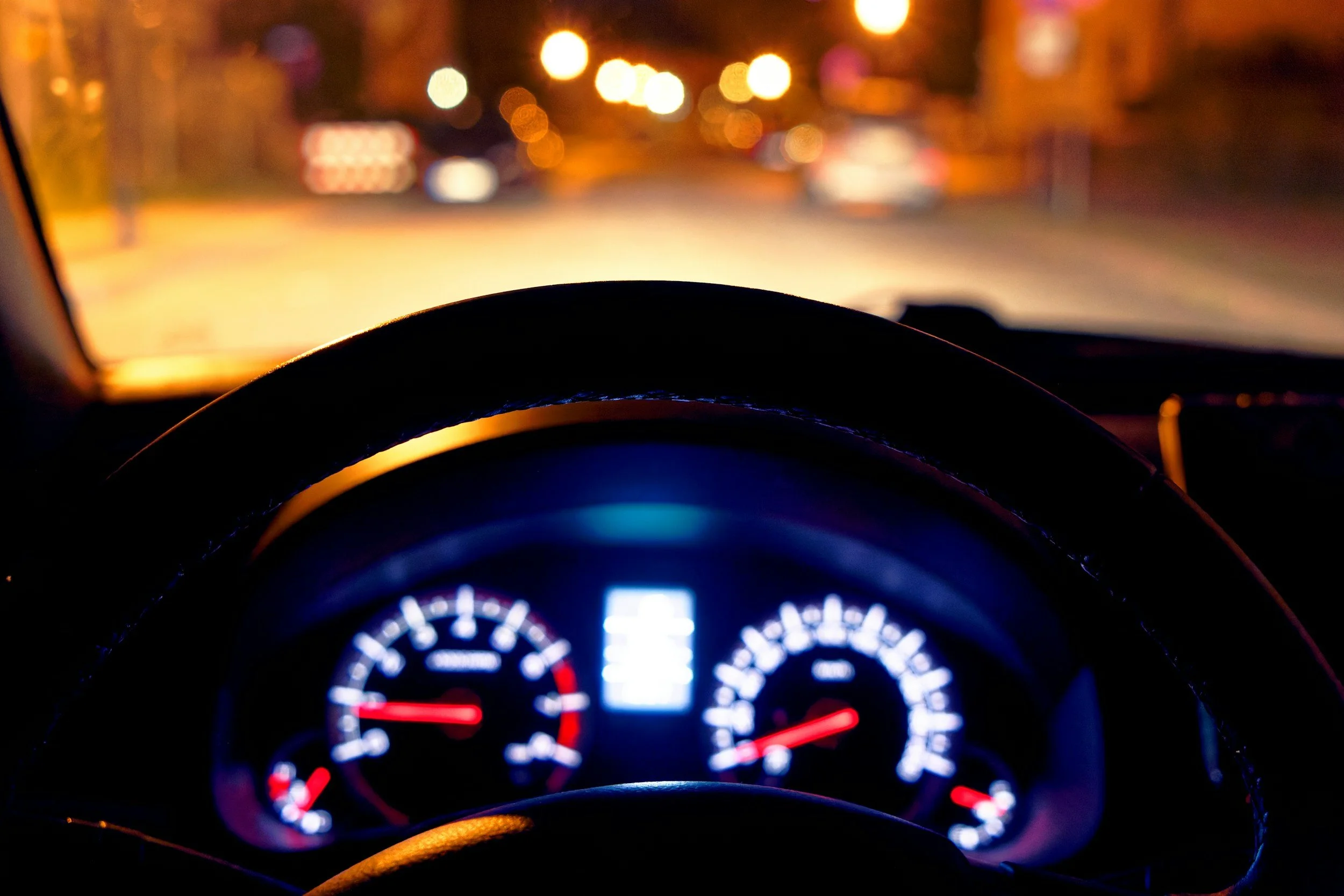 A view from inside a car at night showing the illuminated dashboard with speedometer and fuel gauge, and blurred city lights visible through the windshield, an effect from astigmatism induced by a pterygium