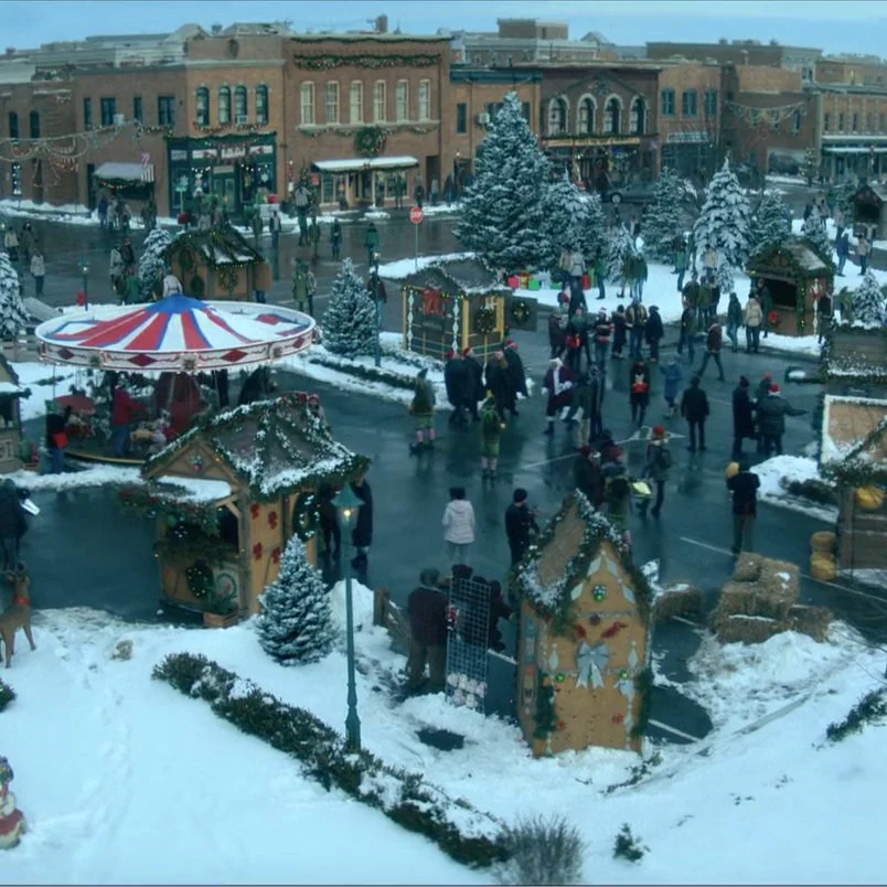 A festive Christmas market scene in a snowy outdoor town square with people shopping and walking among decorated booths, Christmas trees, and holiday ornaments.