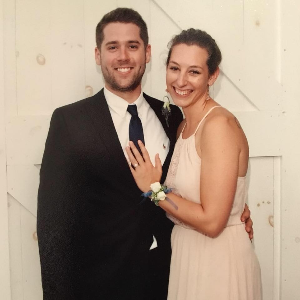 A smiling couple dressed in formal attire, with the woman showing an engagement ring on her finger, standing against a white wooden background.