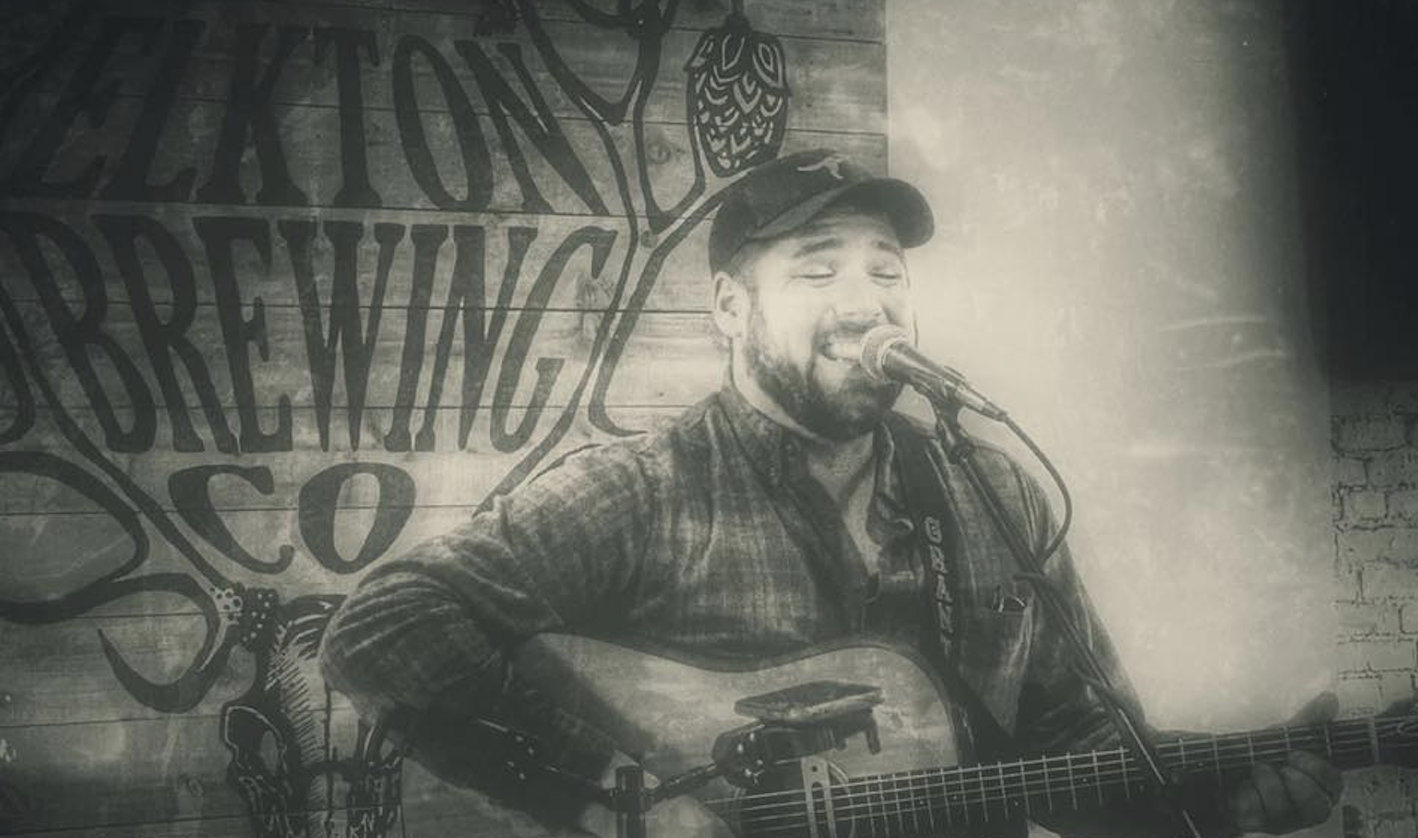 Black and white photo of a man singing into a microphone while playing an acoustic guitar, in front of a wooden wall with a large mural reading "Celebration Brewing Co." and a small illustration of an owl.