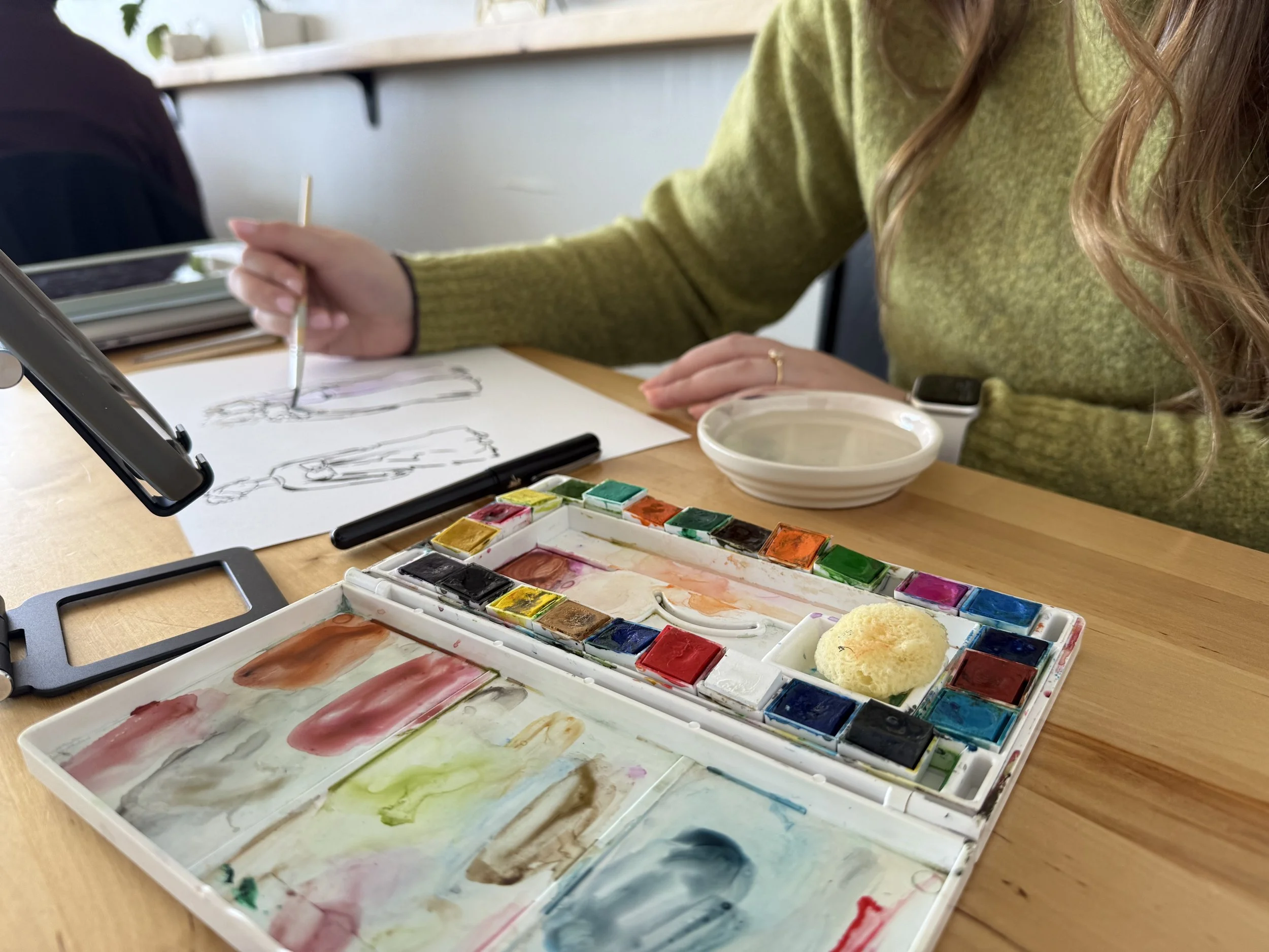 A person is painting sketches of figures with watercolor paints on paper, with a palette of watercolor paints on the table.
