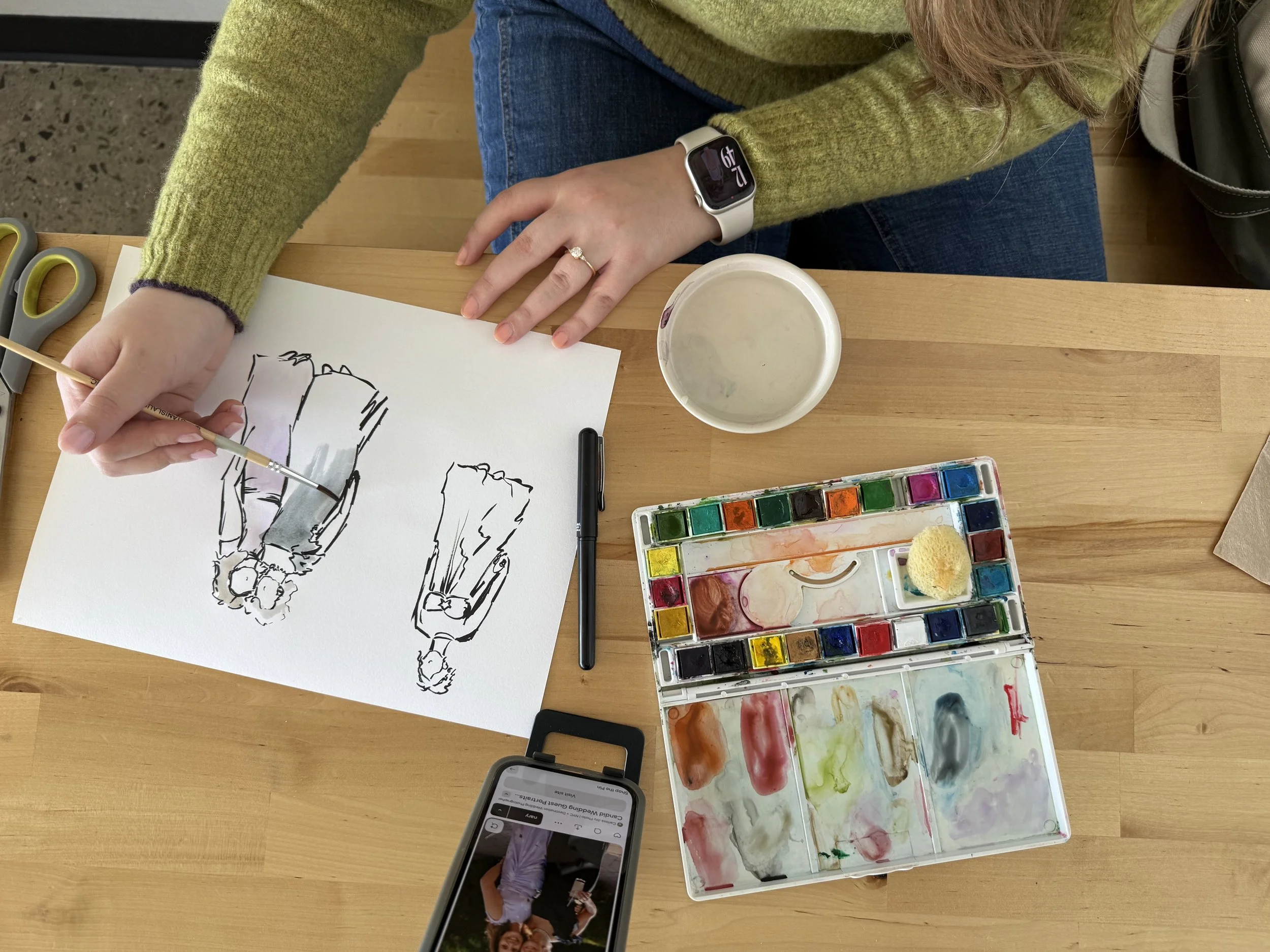 A person painting watercolor illustrations of fashion models on a white sheet of paper, using a fine brush, with watercolor paints and brushes on a wooden table.