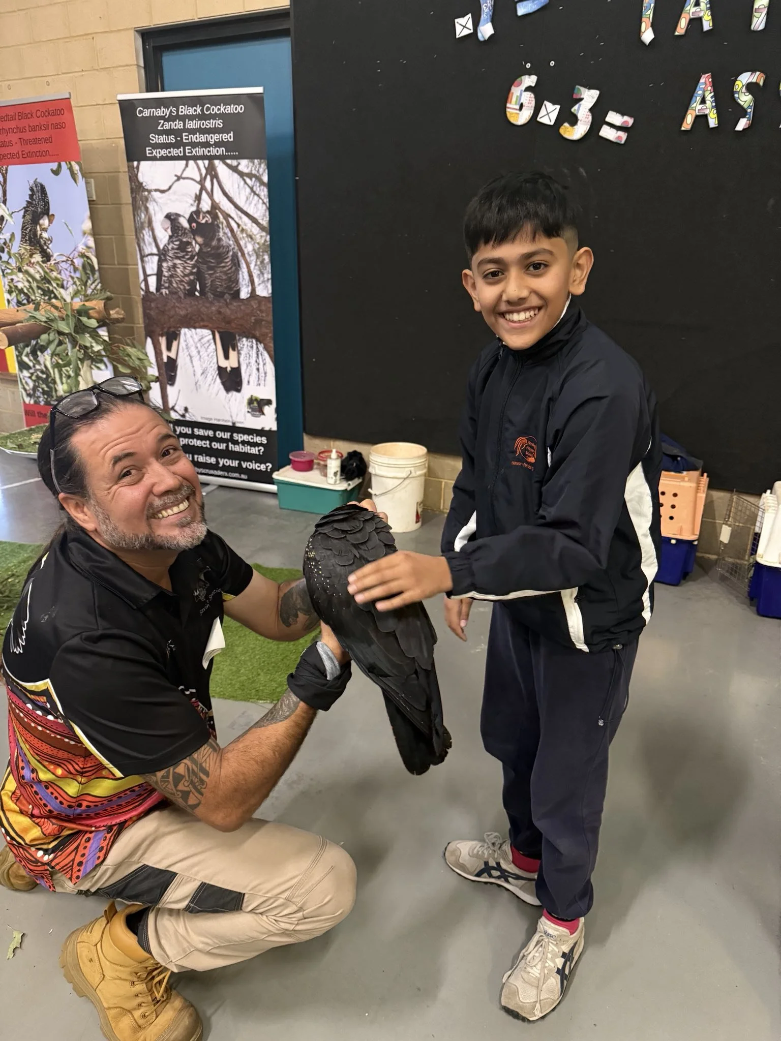 A boy smiling and touching a black bird held by a man with tattoos indoors, with educational posters about the black cockatoo in the background.