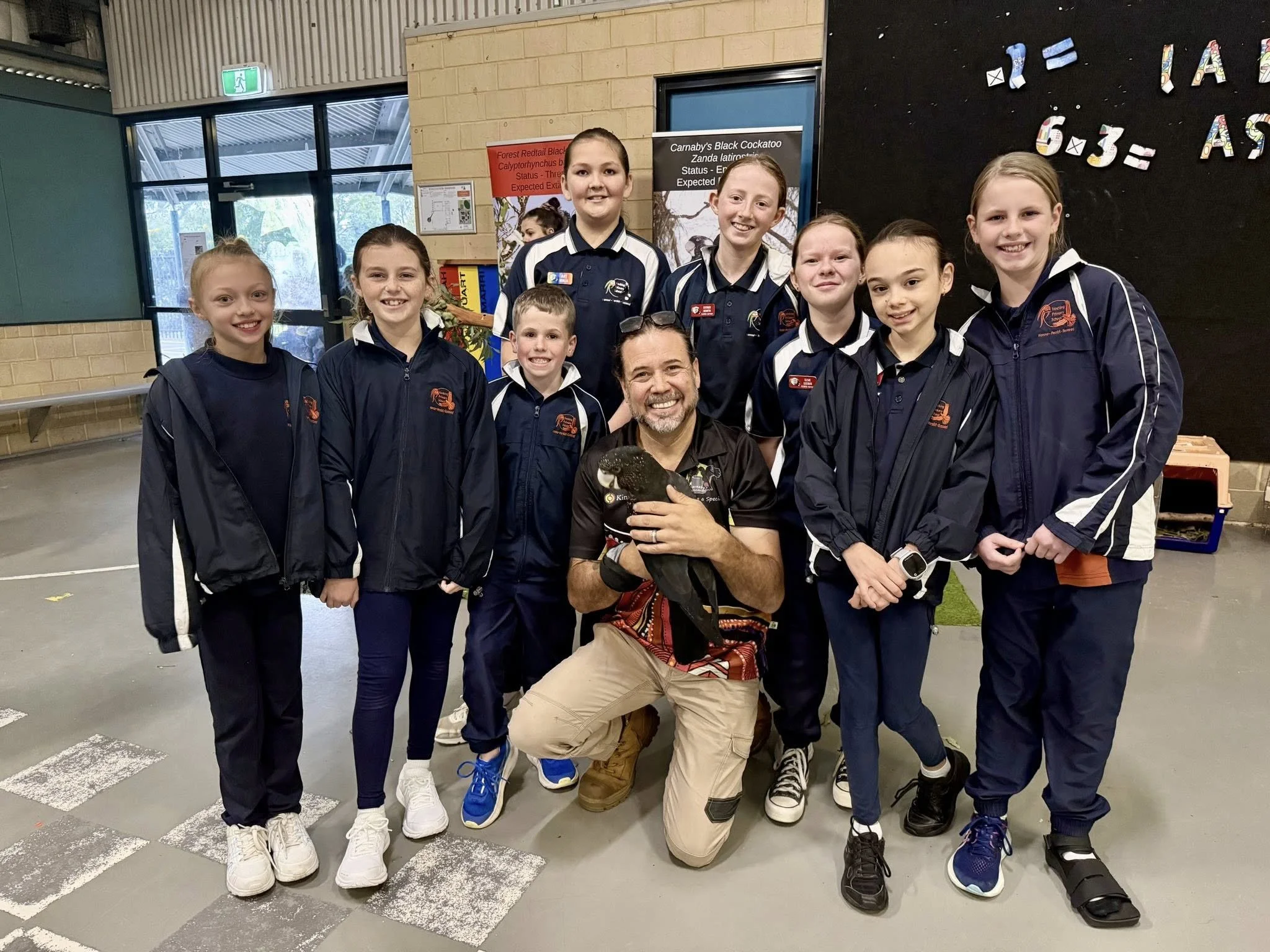 A group of young girls and a man posing inside a building, with the man holding a black bird. The girls are wearing school uniforms and jackets, and are smiling at the camera.