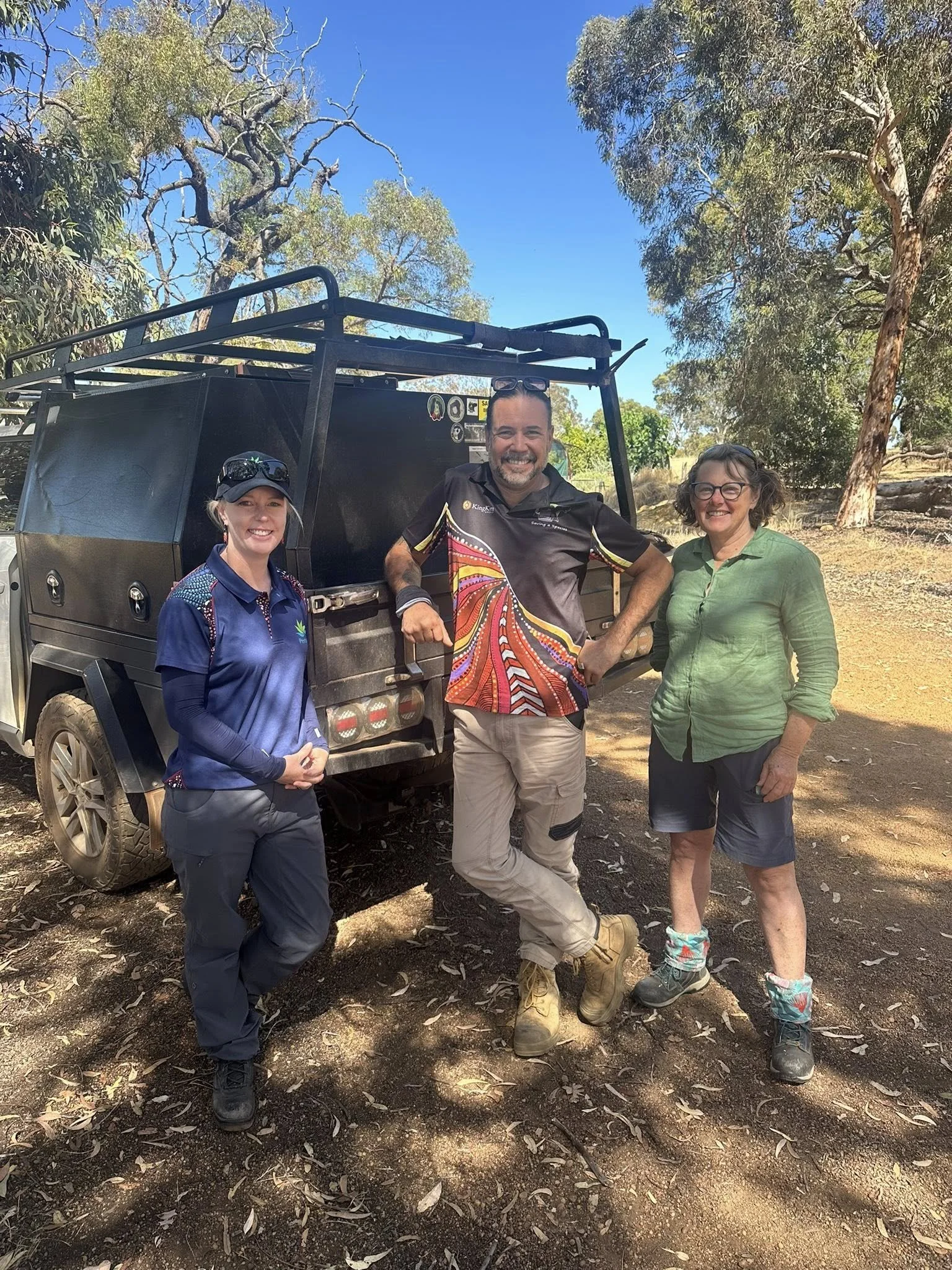 Three people standing outdoors in front of a black utility vehicle, with trees and clear blue sky in the background. They are smiling and dressed in outdoor attire.