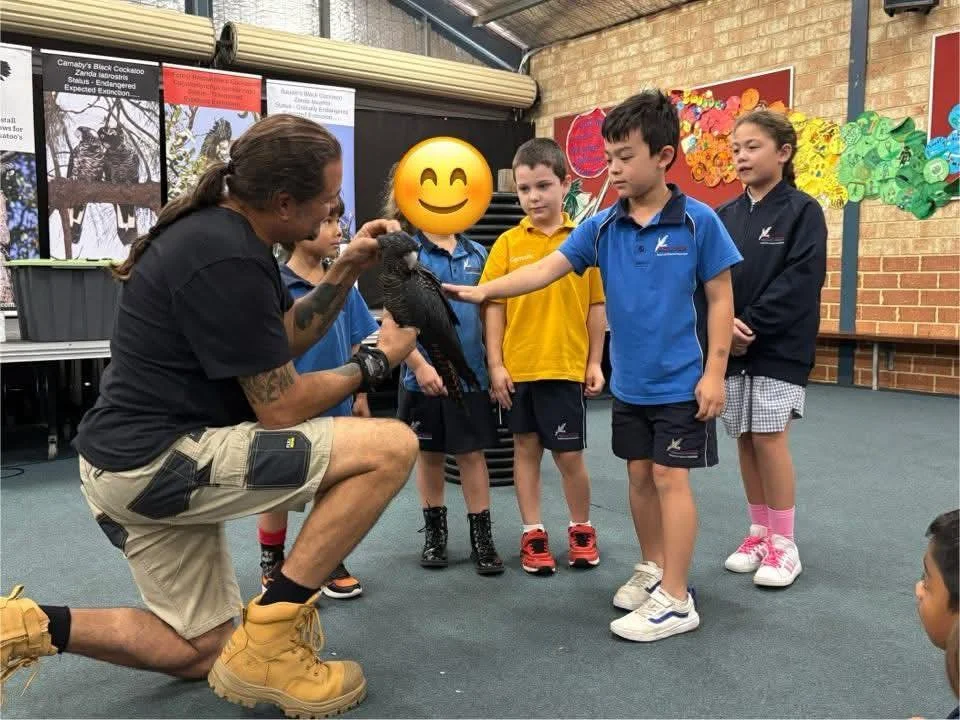 A man with long hair, tattoos on his arm, wearing a black shirt and beige shorts, crouched down holding a large black bird with a yellow face surrounded by children in a classroom or exhibition setting.