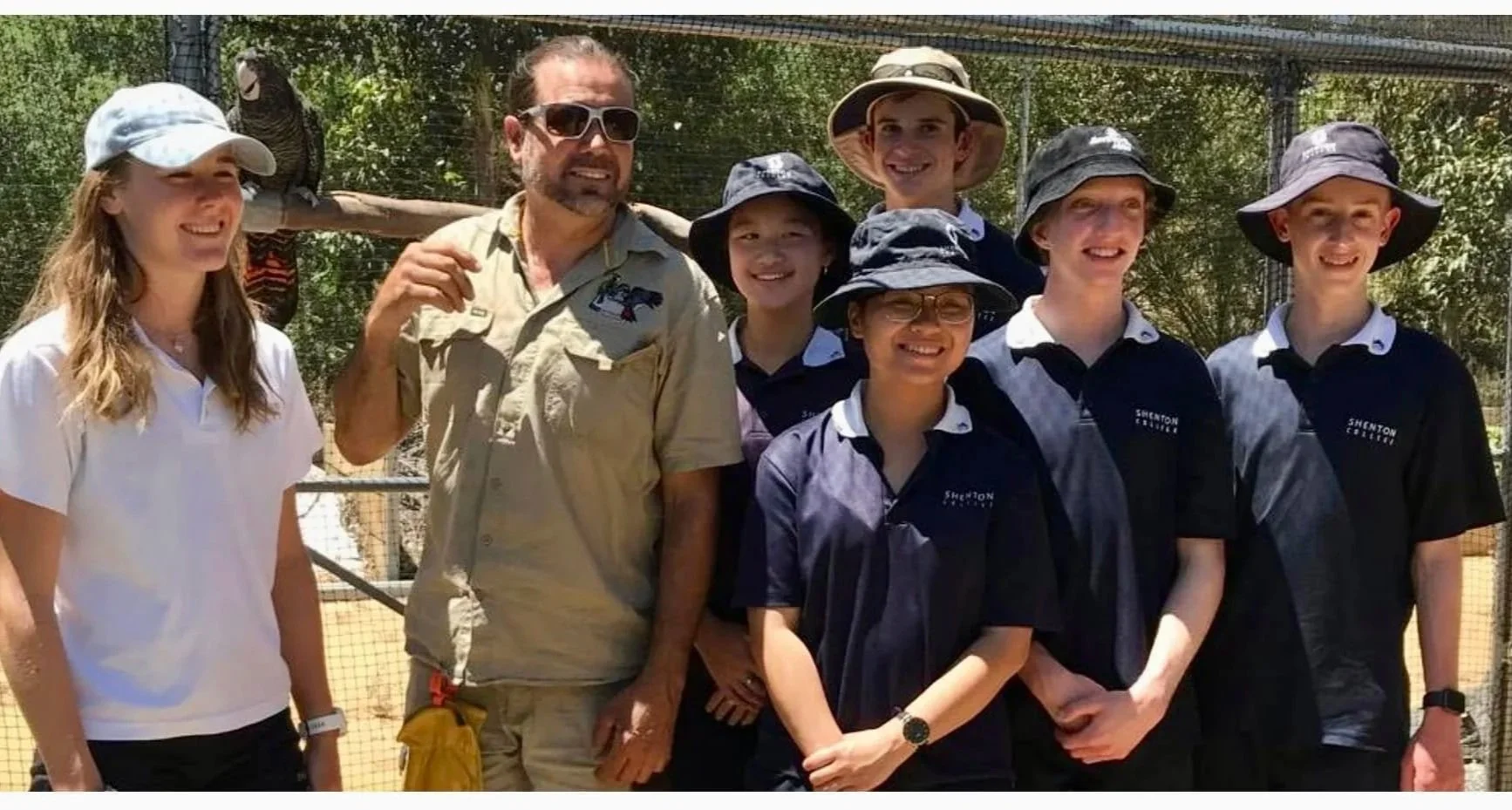 Group of seven people, including children and adults, posing outdoors at a zoo or wildlife sanctuary, with a bird perched on a wooden branch behind them.