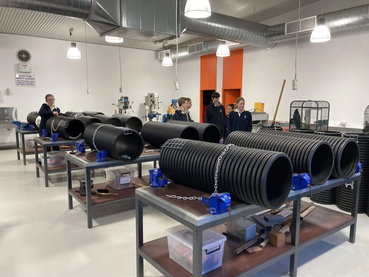 Students in a workshop room working with large black plastic pipes secured with chains and blue clamps, with lab equipment and tools in the background.