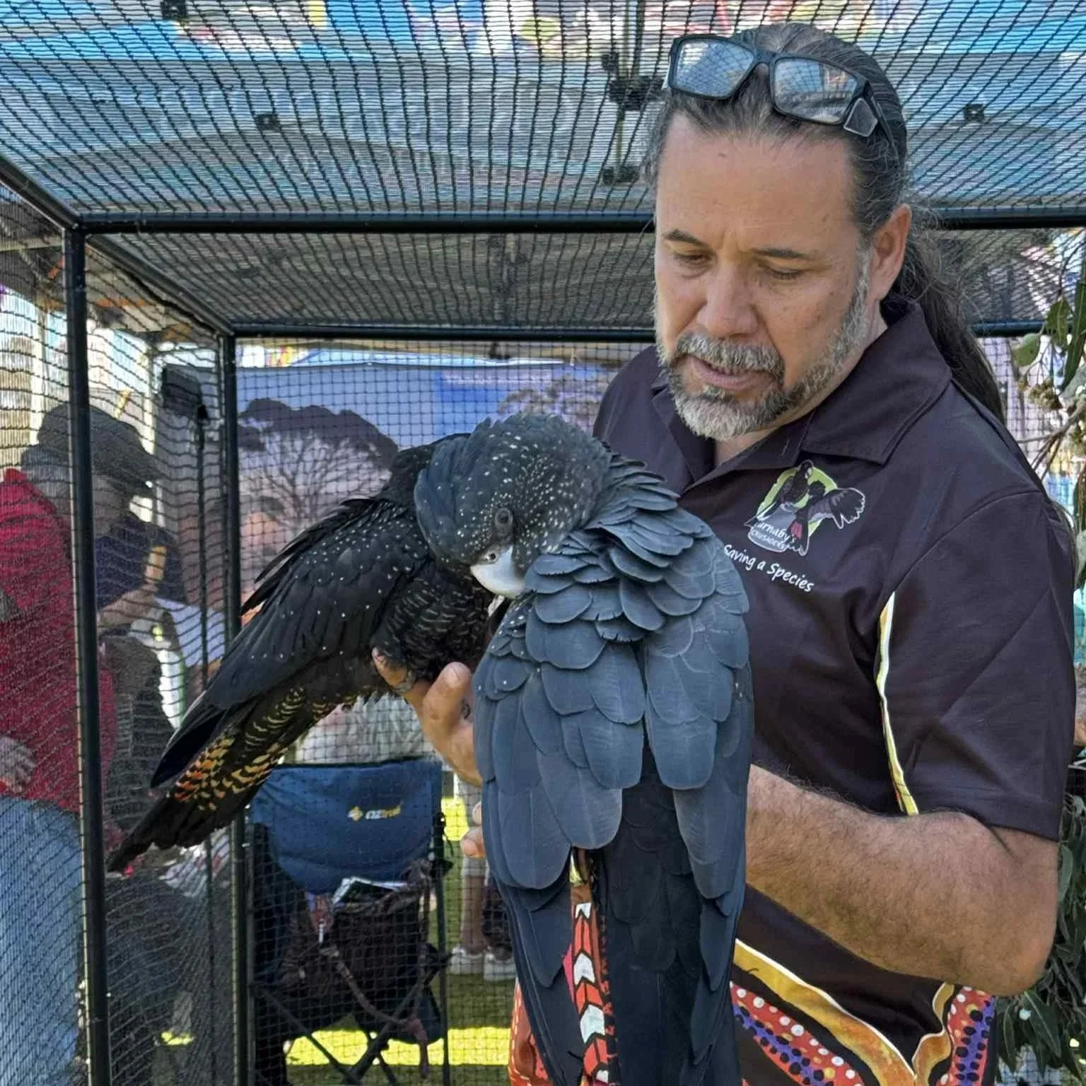 A man holding a black bird with white spots inside a cage at an outdoor event.