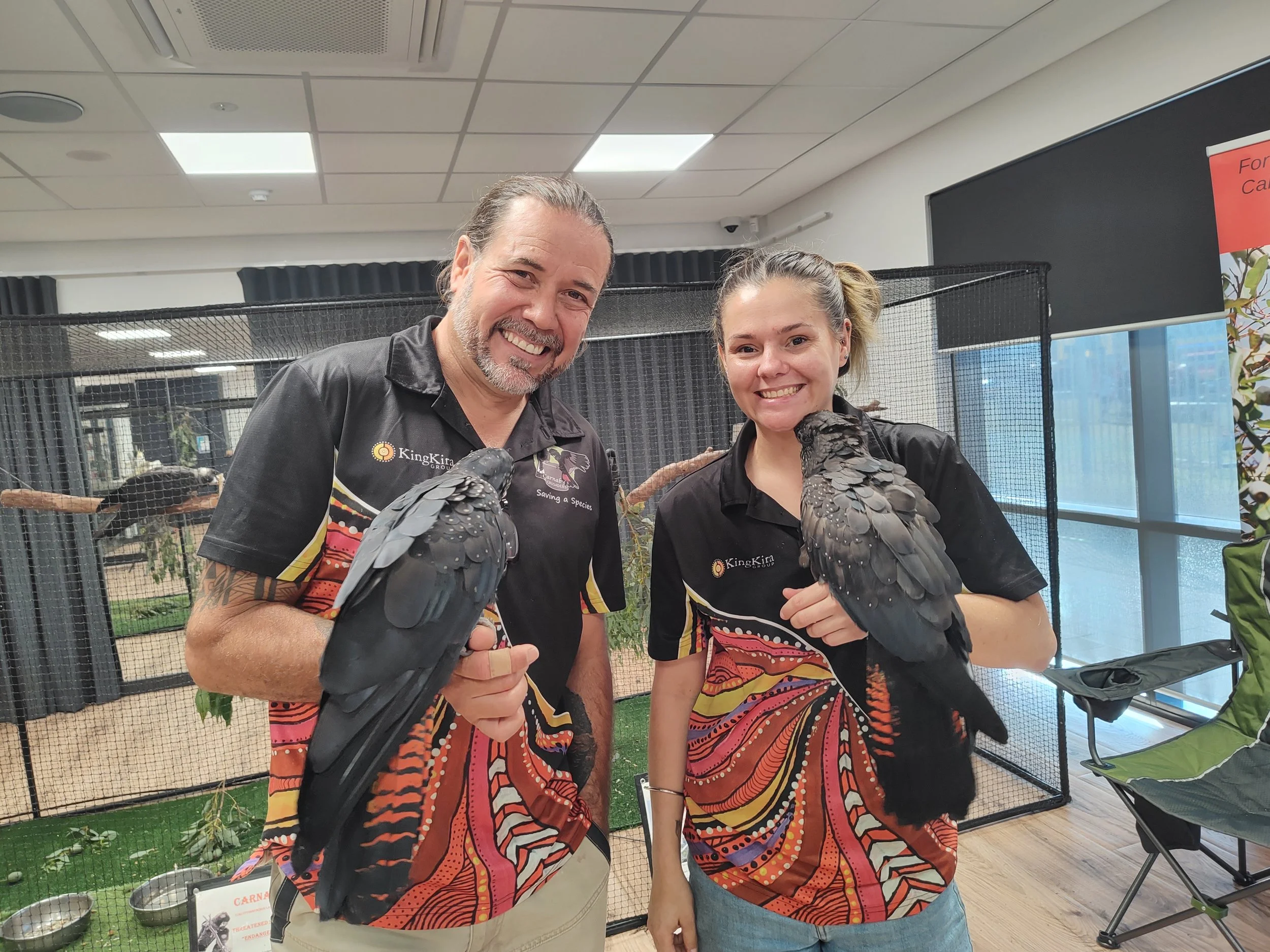 Two people, a man and a woman, smiling and holding black parrots on their arms at an indoor bird exhibit. Both are wearing colorful butterfly-themed shirts.