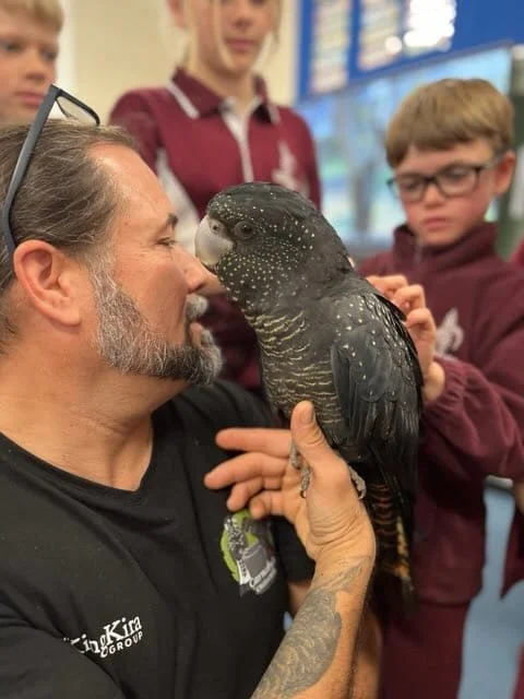 A man holding a black and gray parrot with white spots on its face in a classroom with children observing.