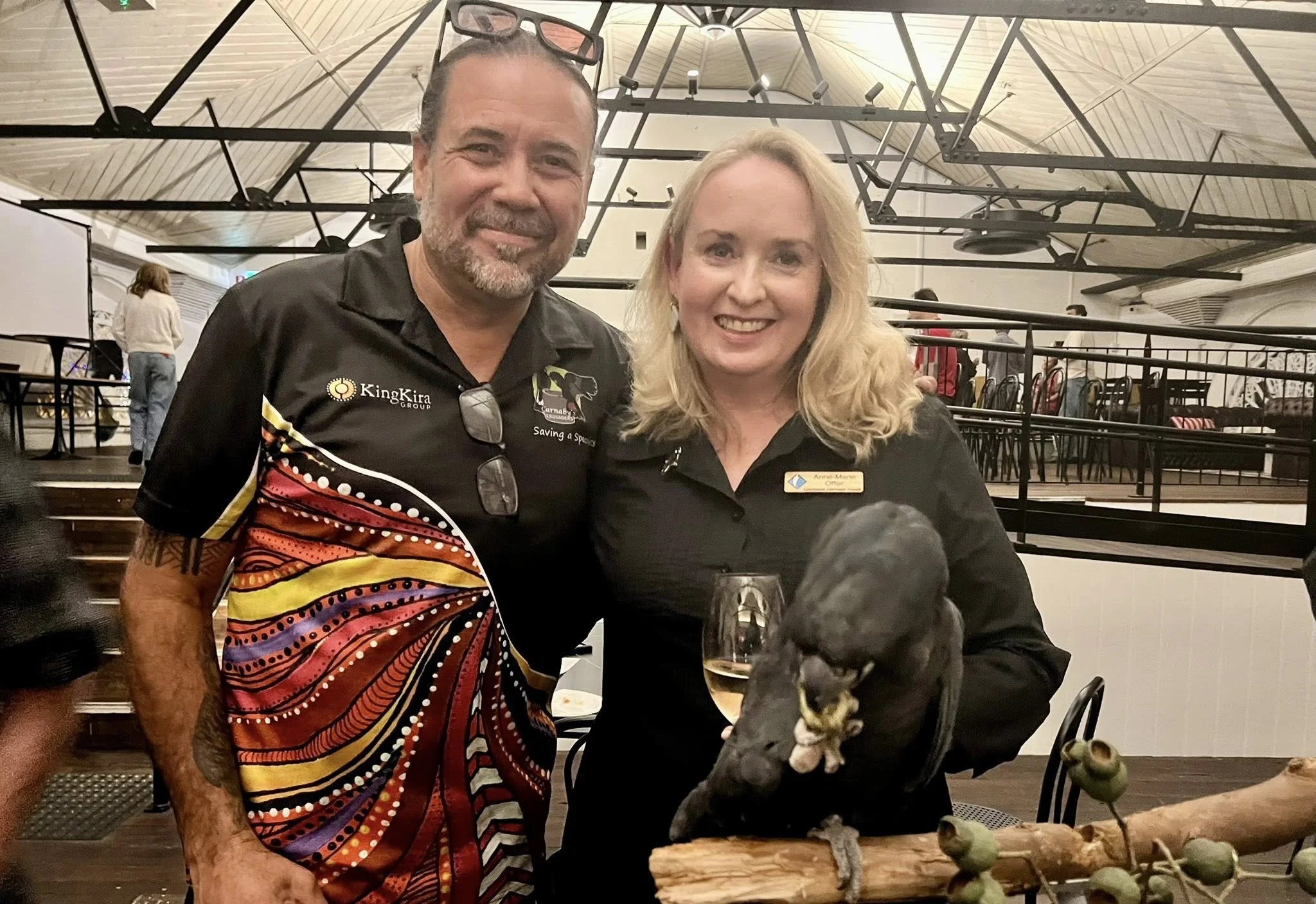 Two smiling people, a man and a woman, are posing together indoors. The man is wearing a black shirt with colorful patterns and text that reads 'KingKira Group,' and has glasses hanging around his neck. The woman is wearing a black shirt with a name tag and is holding a glass of white wine. There is a large black bird, possibly a crow or raven, perched on a decorative branch in front of them. In the background, there are tables, chairs, and several other people in a spacious, well-lit room with a high ceiling.