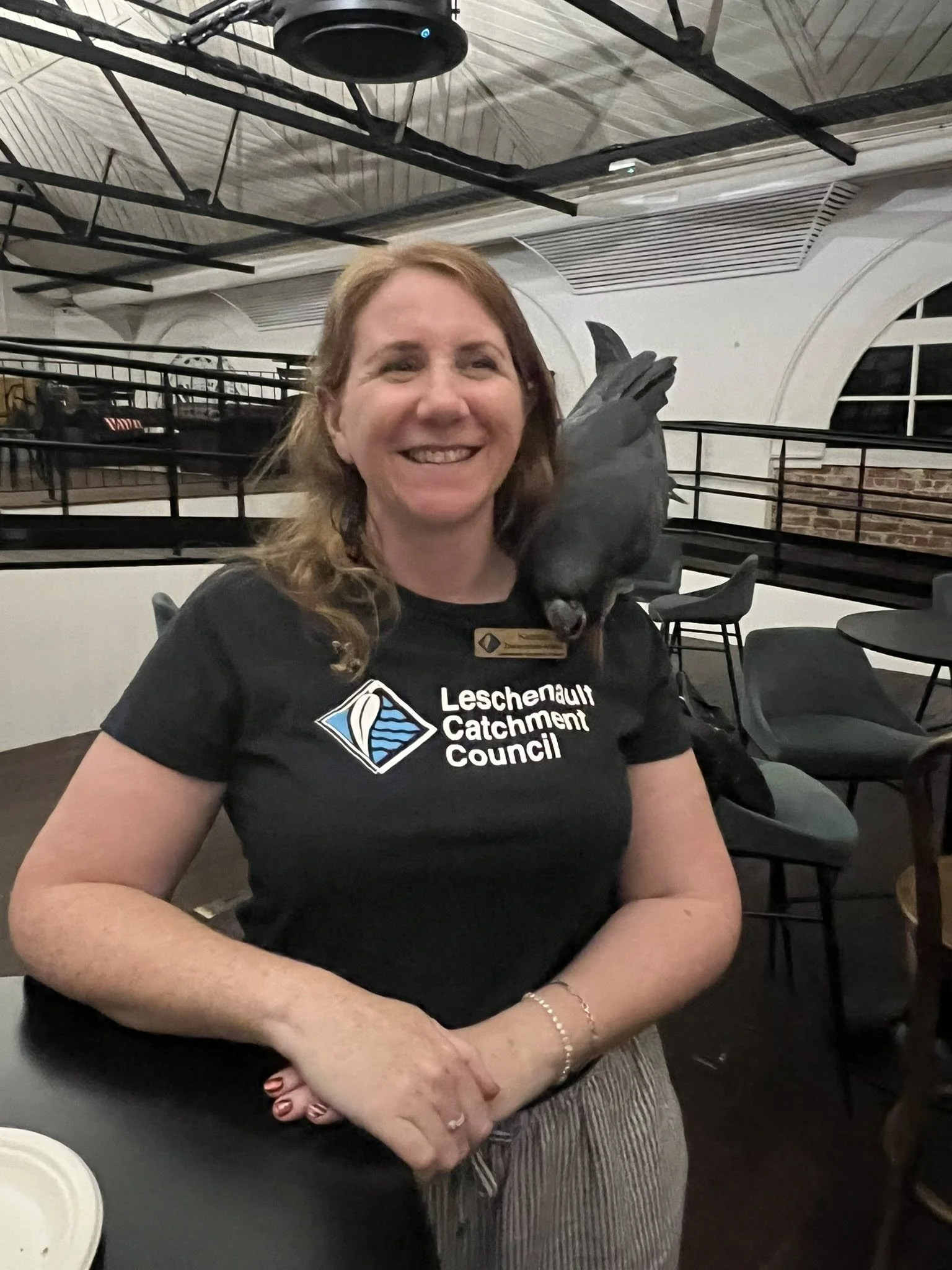 A woman with long red hair smiling, wearing a black Leschenault Catchment Council t-shirt, with a dark-colored bird perched on her shoulder inside a modern indoor space with black chairs and white walls.