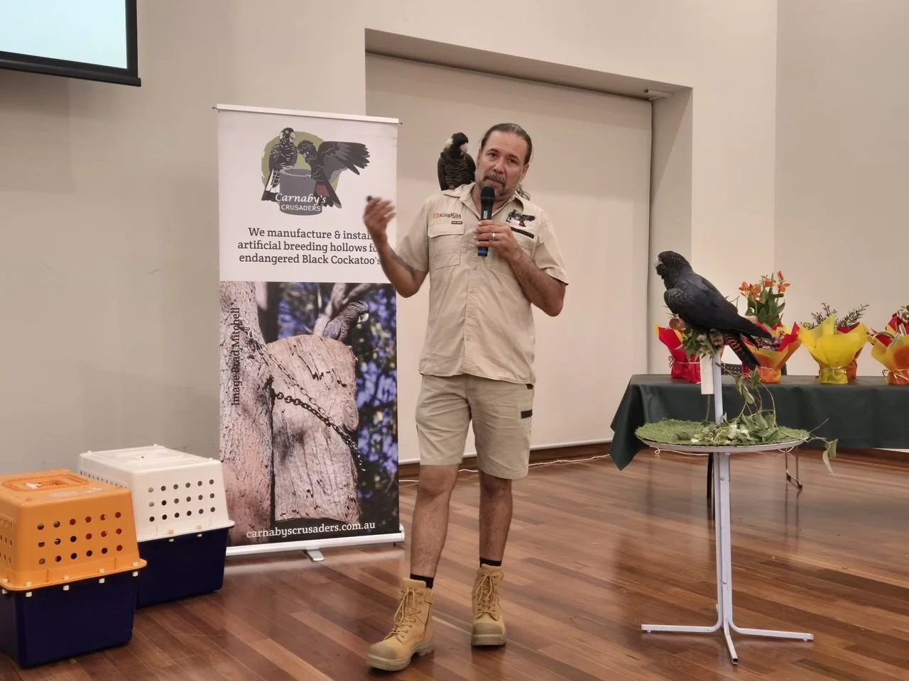 A man in khaki uniform with a black cockatoo perched on his shoulder is speaking into a microphone in a room with wooden flooring. There is a table with another black cockatoo and flower arrangements, and a banner advertising black cockatoo breeding 
