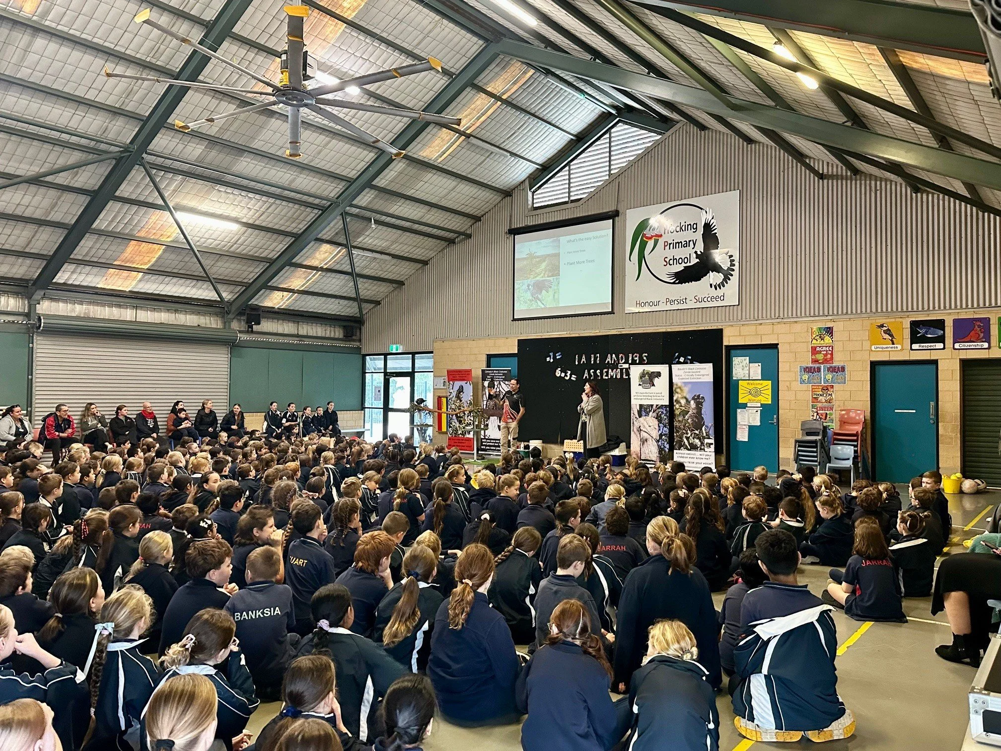 School assembly in a large gymnasium with students seated on the floor and teachers standing at the front. The school is called Nocking Primary School, with a logo and motto 'Honour - Persist - Succeed.' A presentation is displayed on a projector screen. The students are wearing dark uniforms.