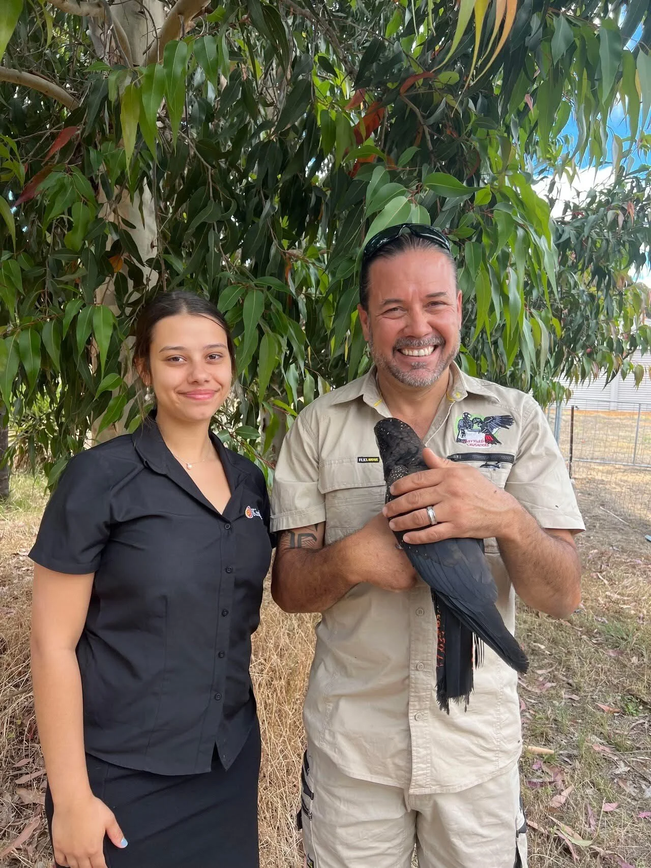 A woman and a man standing outdoors in front of a tree, smiling at the camera. The man is holding a large black bird, possibly a black cockatoo.