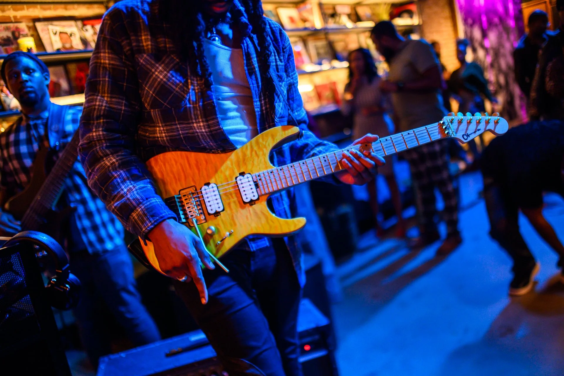 A person playing an electric guitar in a lively, colorful environment with other people in the background.