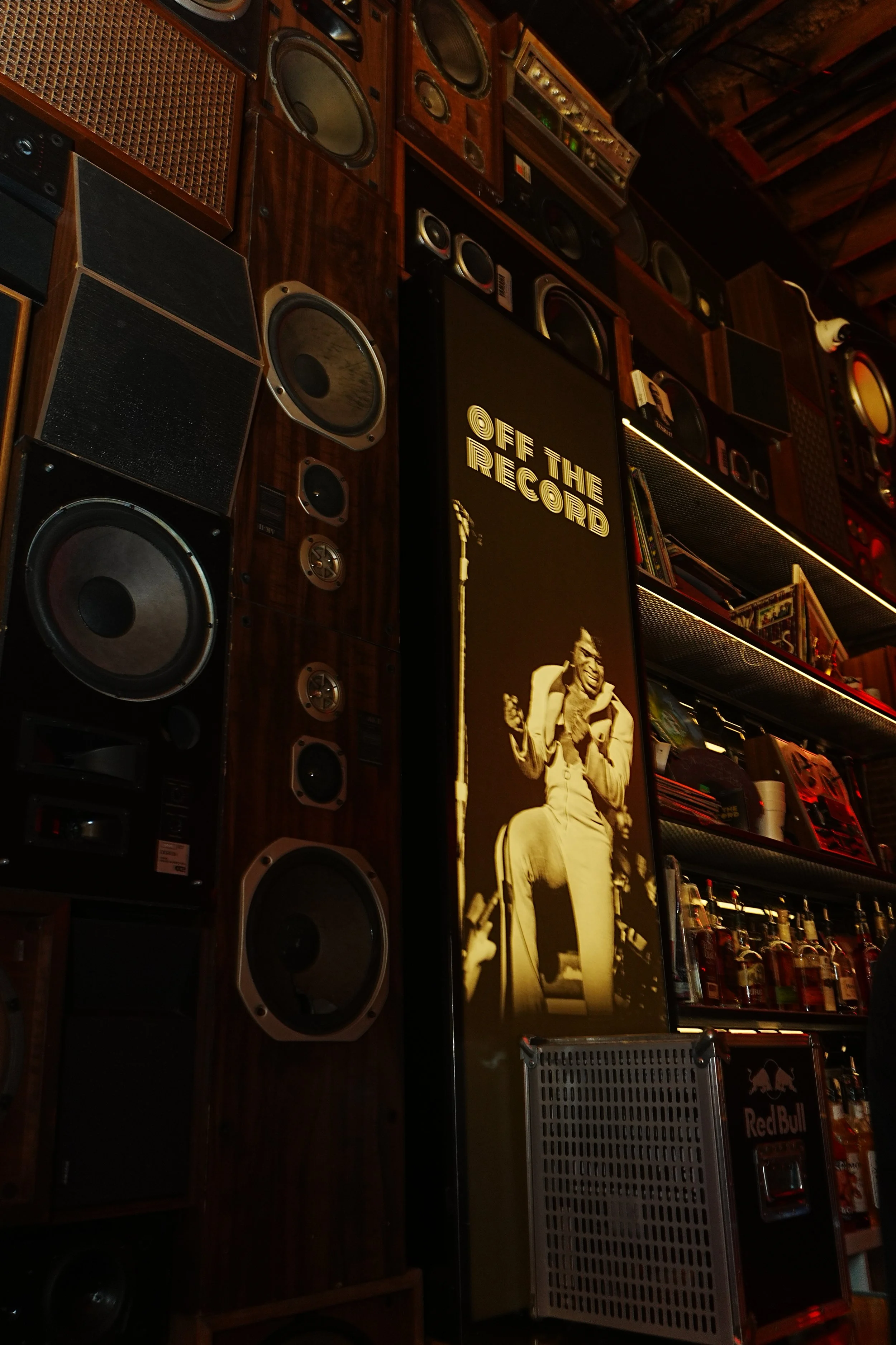 Interior of a room with vintage speakers and audio equipment, a photo of a guitarist with the text 'Off the Record' on a black background, and shelves stocked with liquor bottles.