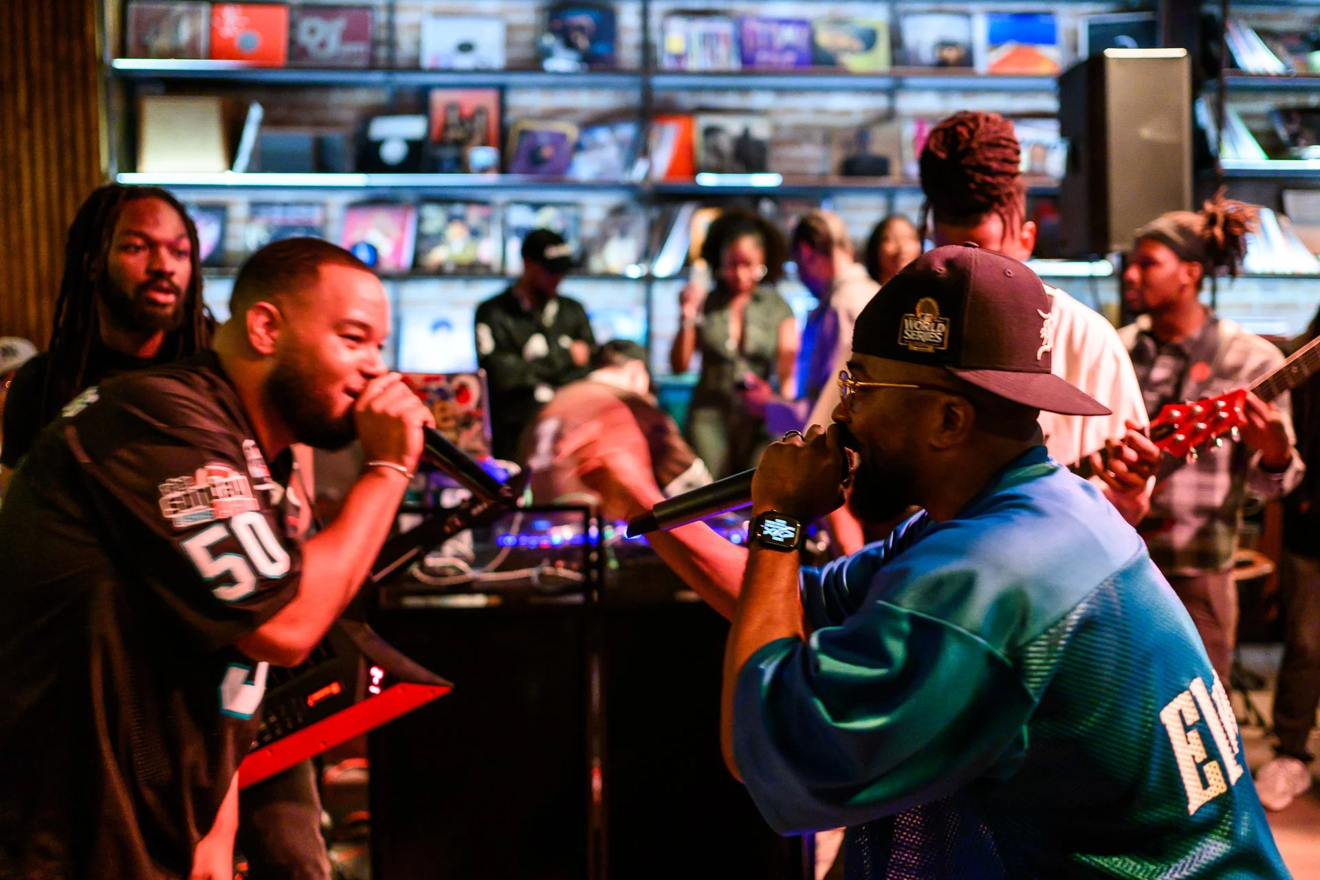 A group of people, including two men in the foreground holding microphones, are performing karaoke in a lively, colorful bar with a brick wall and shelves filled with books or records in the background.