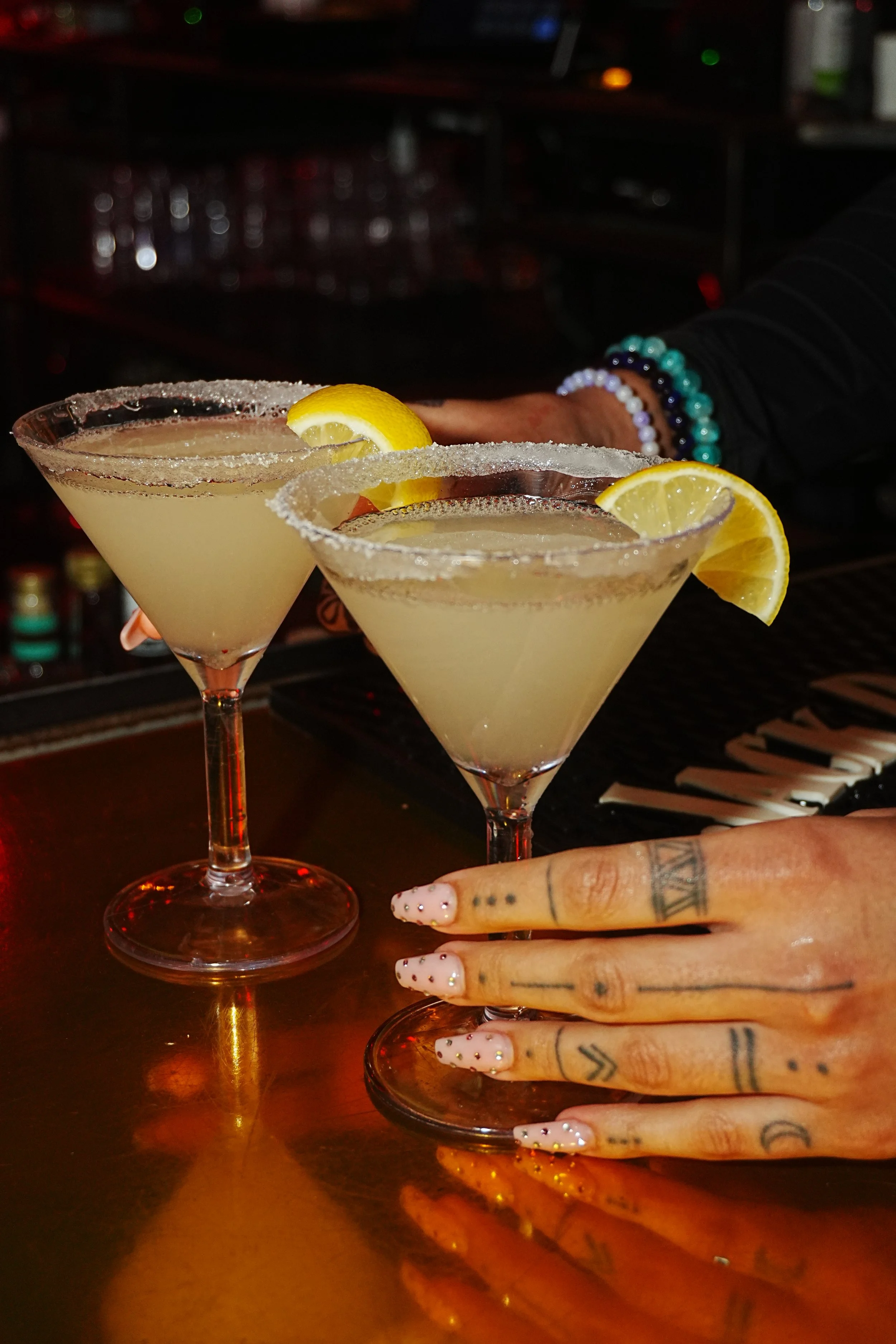 Two lemon margarita cocktails on a bar counter with a person's tattooed hand beside them.