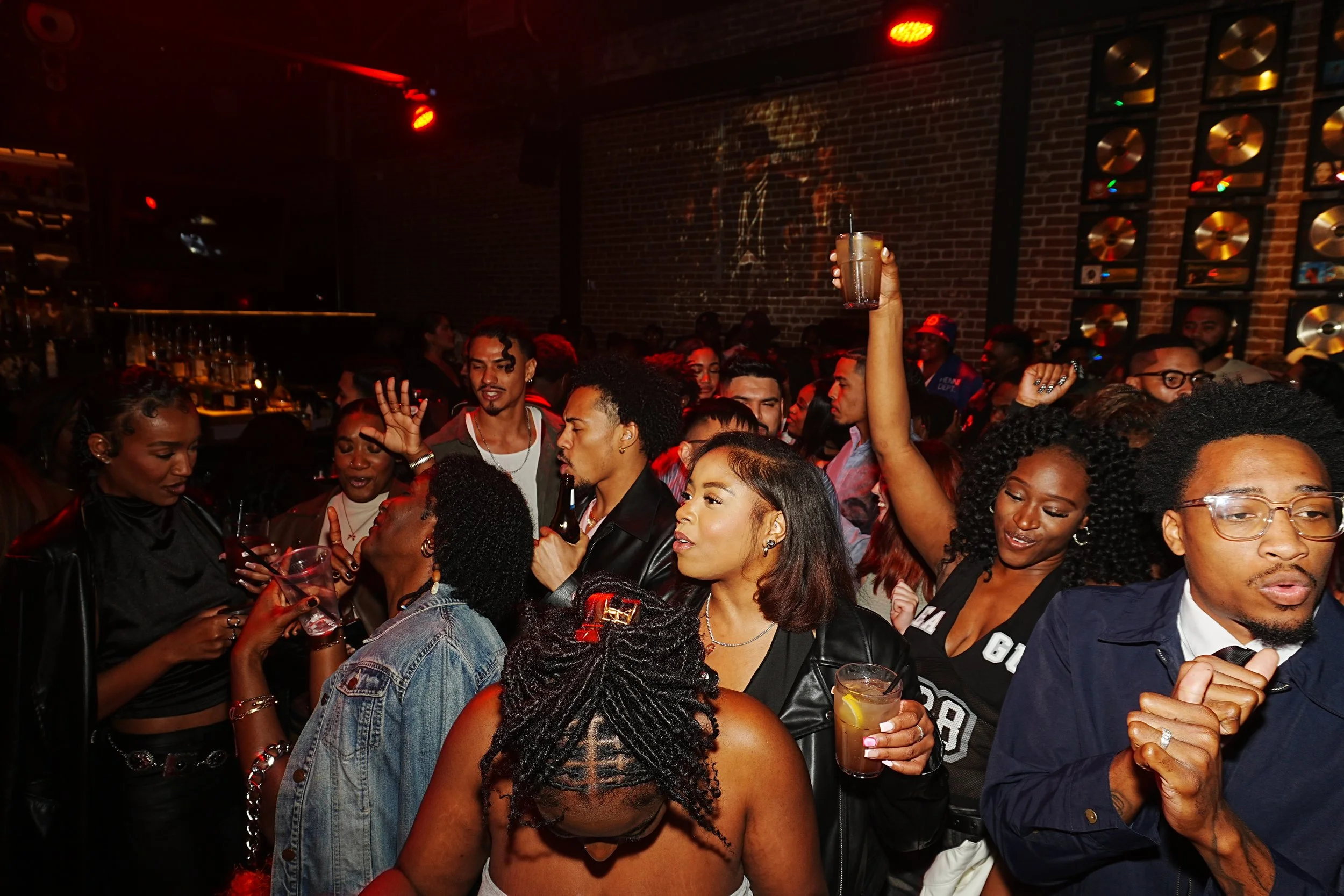 Crowd of young adults enjoying a lively night at a bar or club, with some holding drinks and dancing, against a brick wall with gold records.