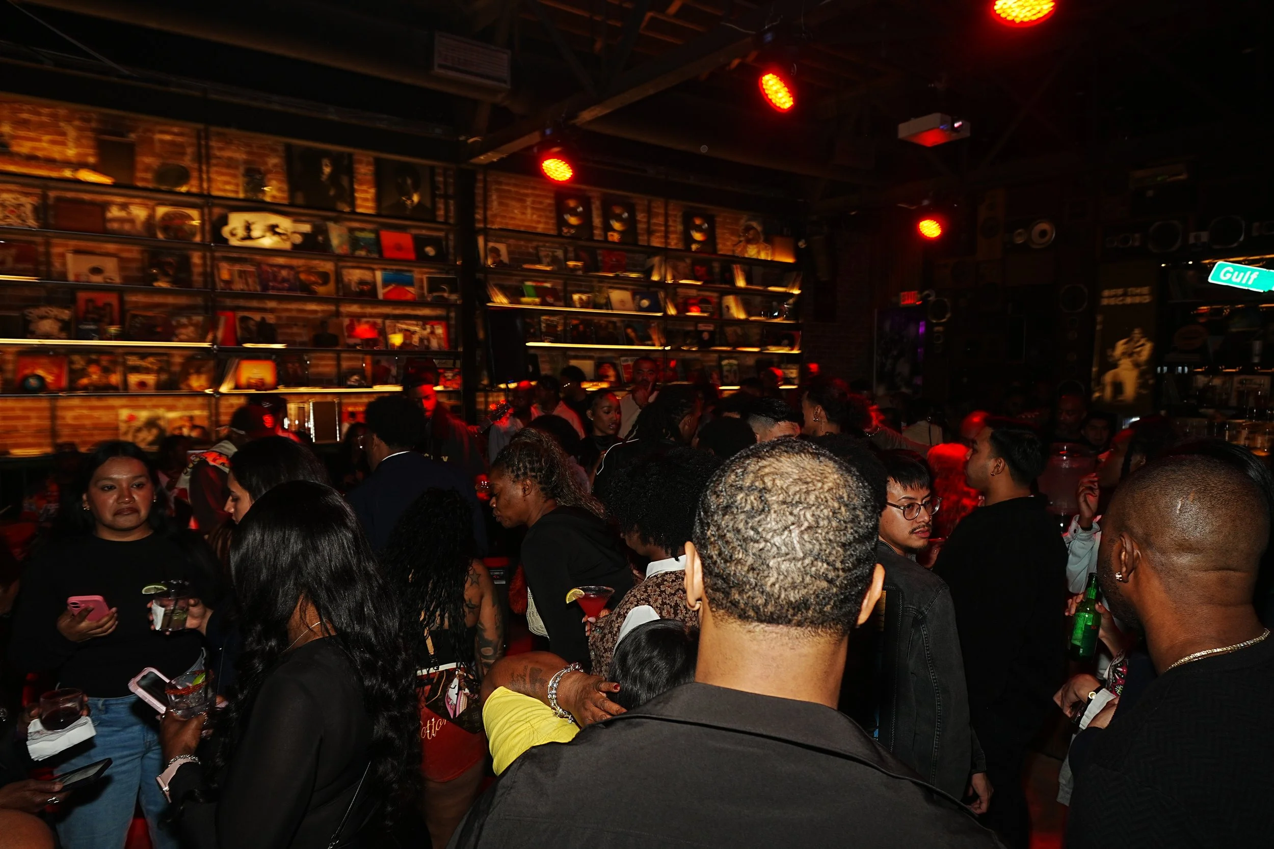 Crowd of people at a bar or nightclub with dim lighting and red spotlights, brick walls, and shelves filled with records and framed pictures.