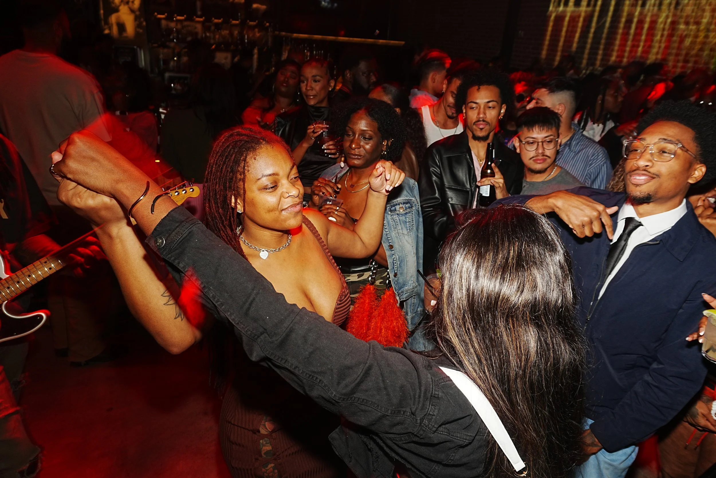 A lively party scene with diverse people dancing and socializing in a dimly lit space, red lights, and a musician playing guitar visible on the left.