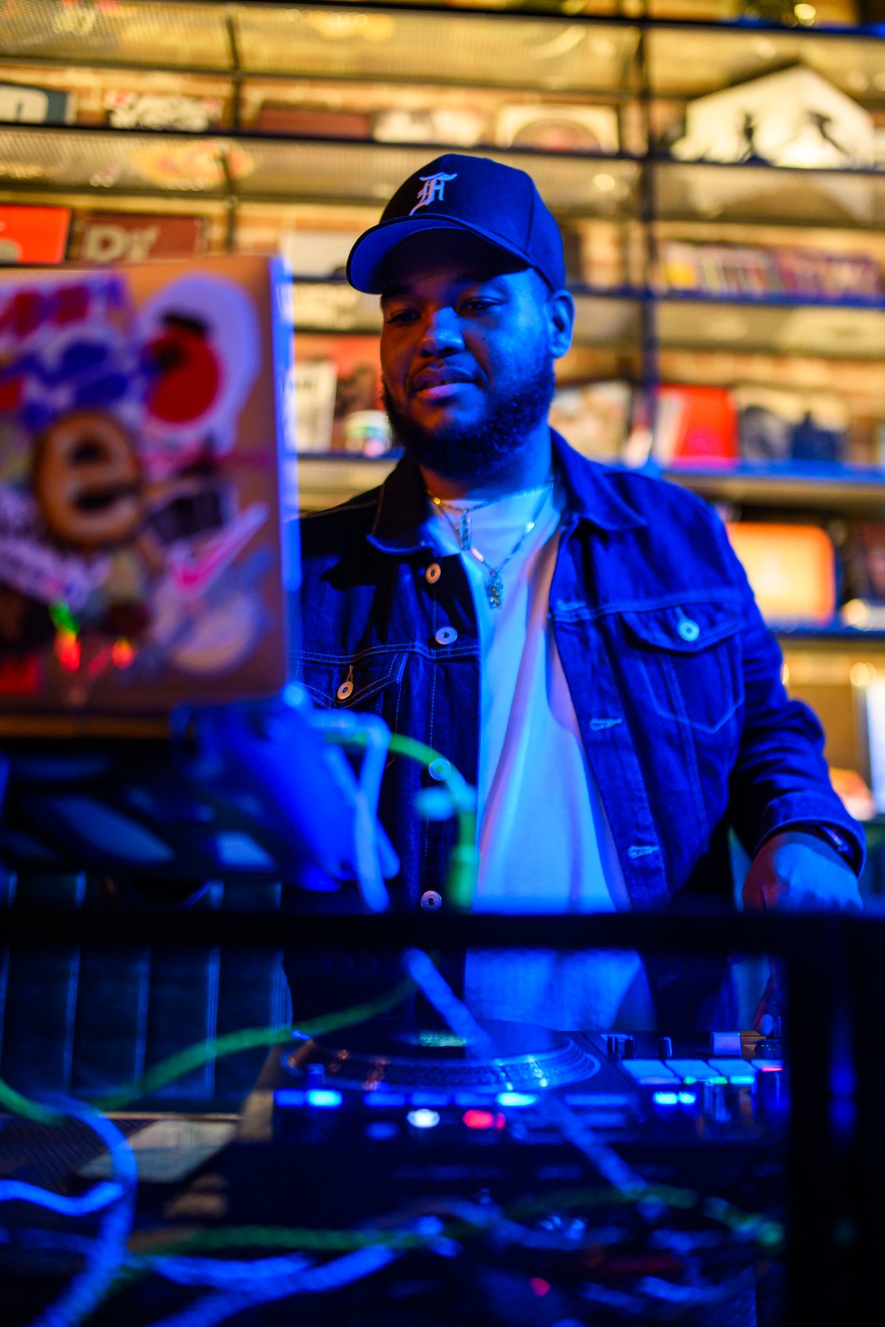 A man djing at a black table with a turntable and laptop, wearing a black baseball cap and denim jacket in a colorful, dimly lit setting.