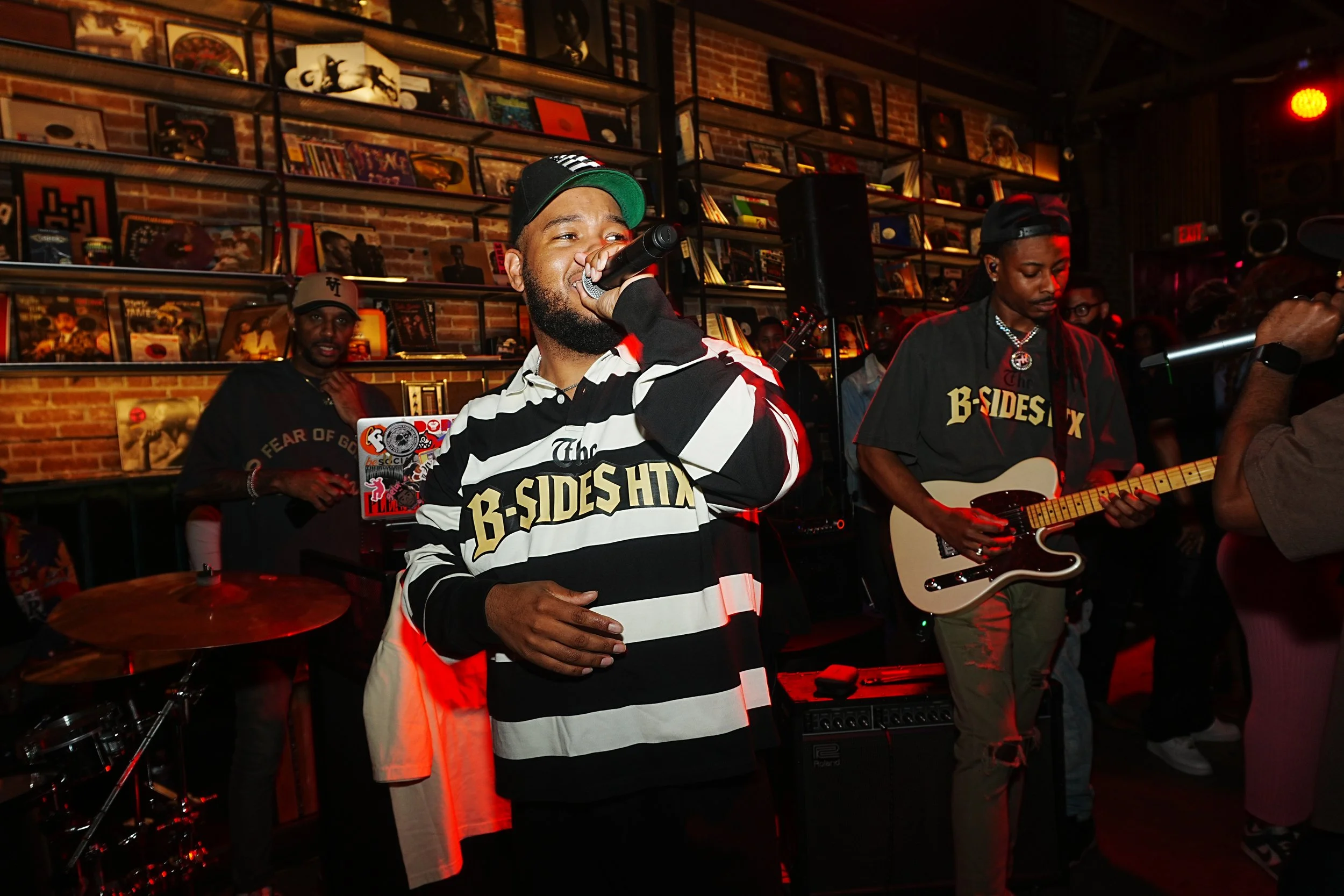 A musician singing into a microphone while another plays guitars in a dimly lit venue with brick walls and shelves of records.