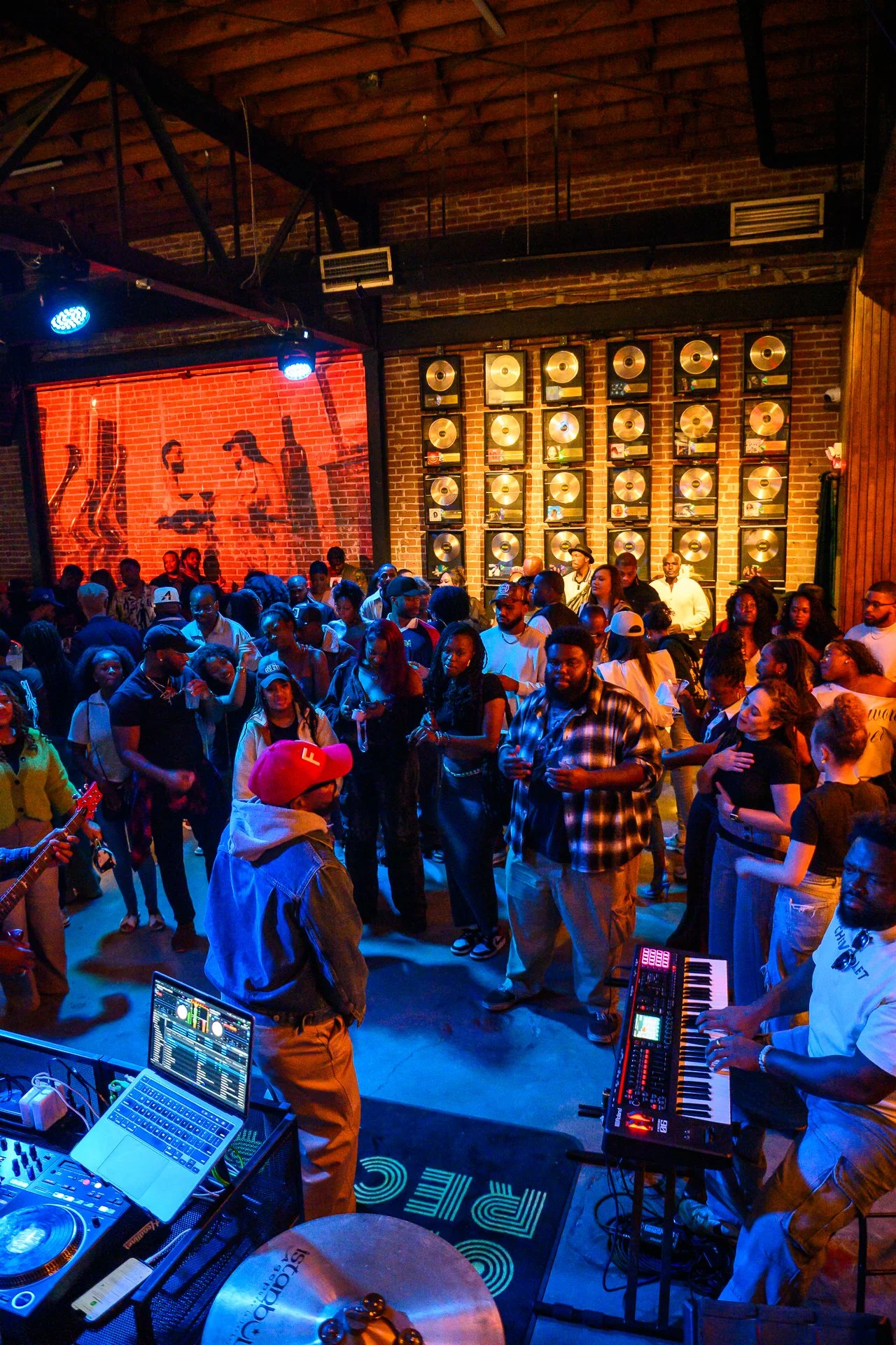 A lively indoor music venue with a crowd of people dancing and socializing. The scene features a DJ setup, musical instruments, and a brick wall decorated with framed gold records. The lighting is colorful, creating a vibrant atmosphere.